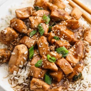 A plate of hoisin chicken served over white rice, topped with sesame seeds and chopped green onions. Two chopsticks are placed beside the dish.