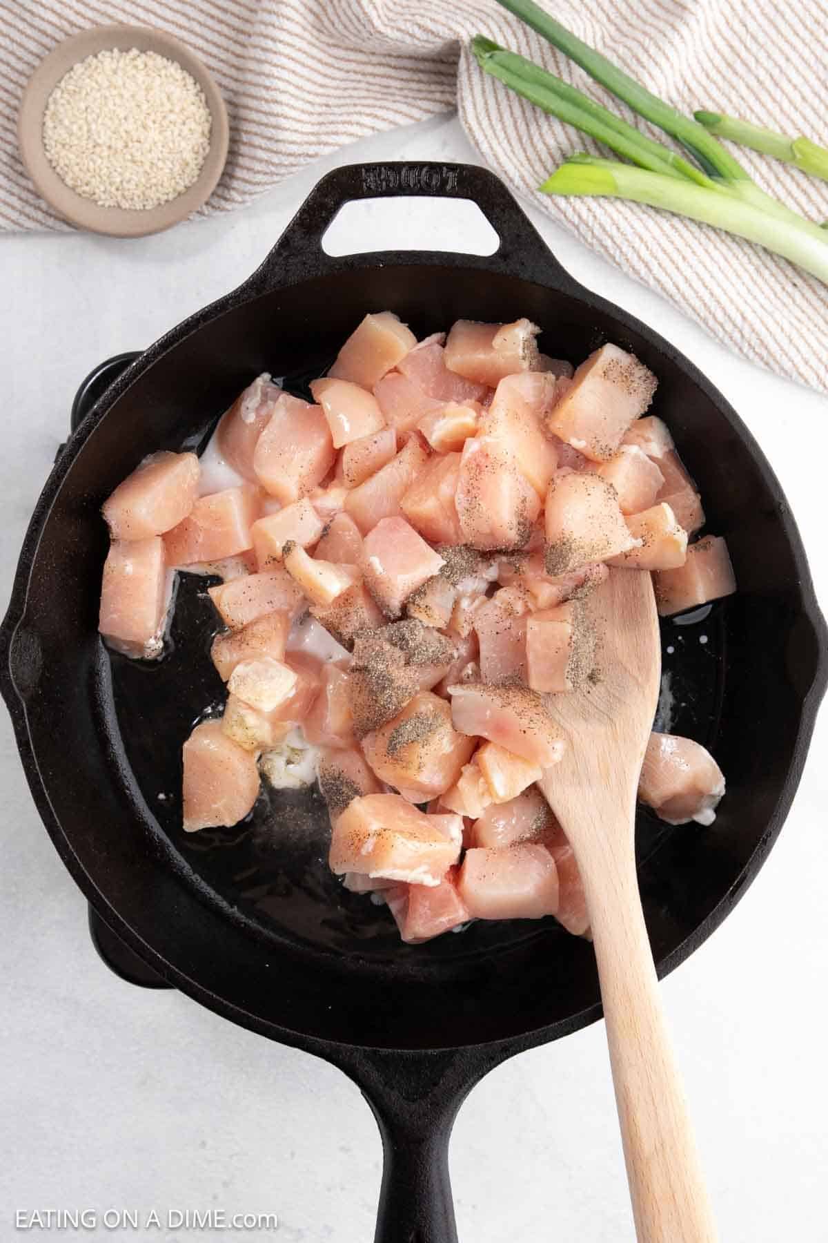 Diced chicken pieces being sautéed in a cast iron skillet with a wooden spoon, seasoned with pepper. Nearby, green onions and a dish of sesame seeds await their turn to elevate this Hoisin Chicken dish on a light surface.