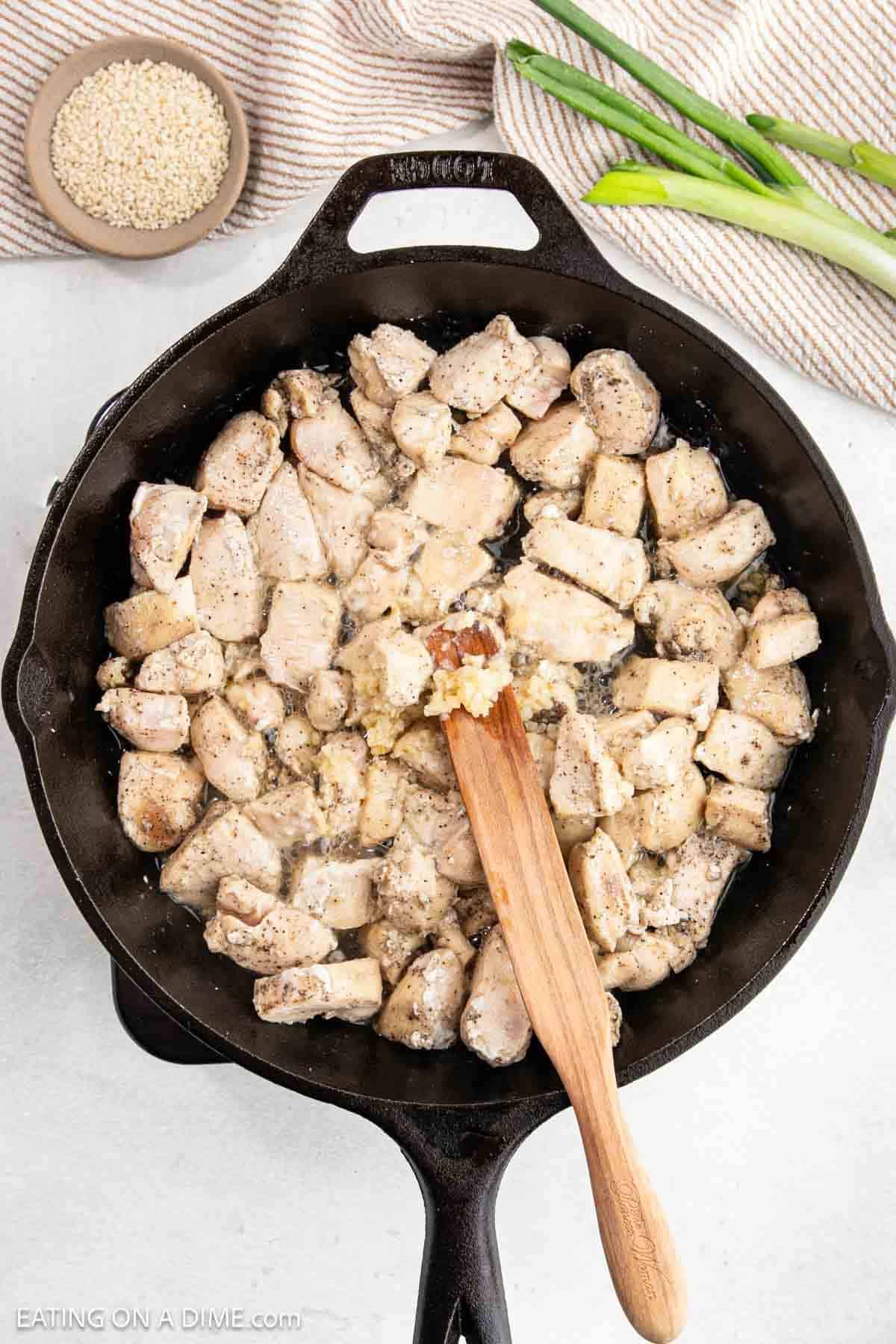 A cast iron skillet filled with hoisin chicken pieces sizzles as they are stirred with a wooden spatula. Uncooked rice and green onions spread across the light surface in the background, waiting to join the savory feast.