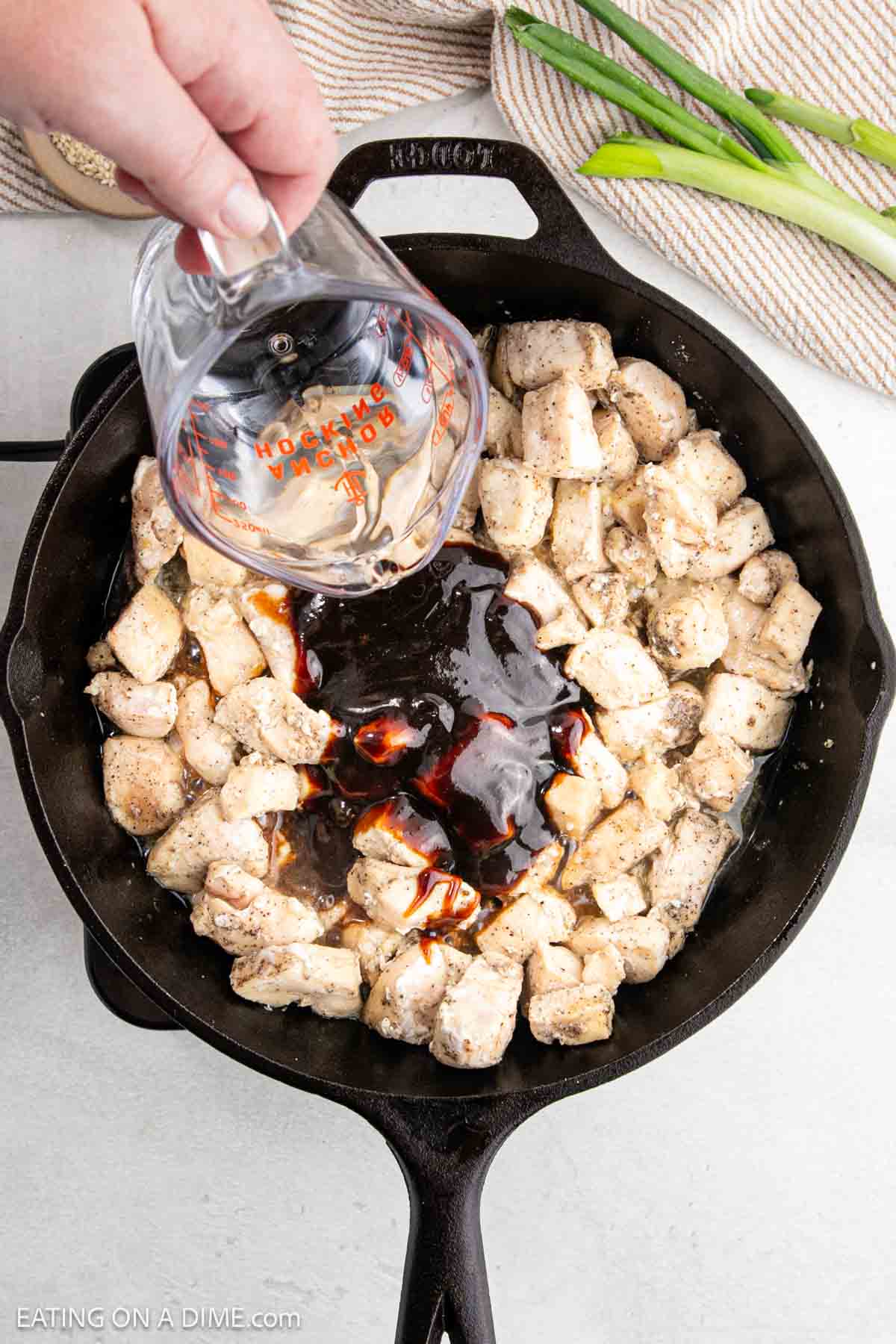 A hand pours teriyaki sauce from a measuring cup into a cast iron skillet filled with diced, cooked hoisin chicken. Green onions and a striped cloth are visible in the background.