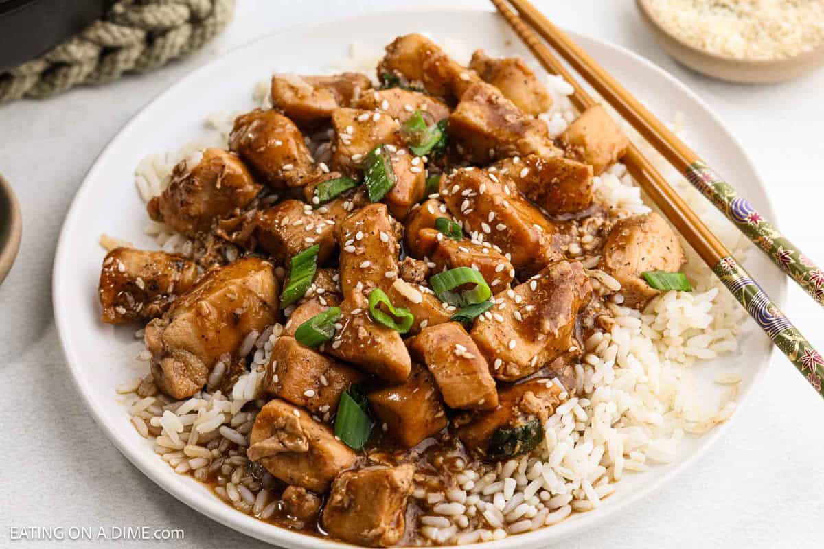 A plate of teriyaki hoisin chicken with chunks of tender meat in a glossy sauce, topped with sesame seeds and chopped green onions over white rice. Chopsticks rest on the side, ready to dive into this savory delight.