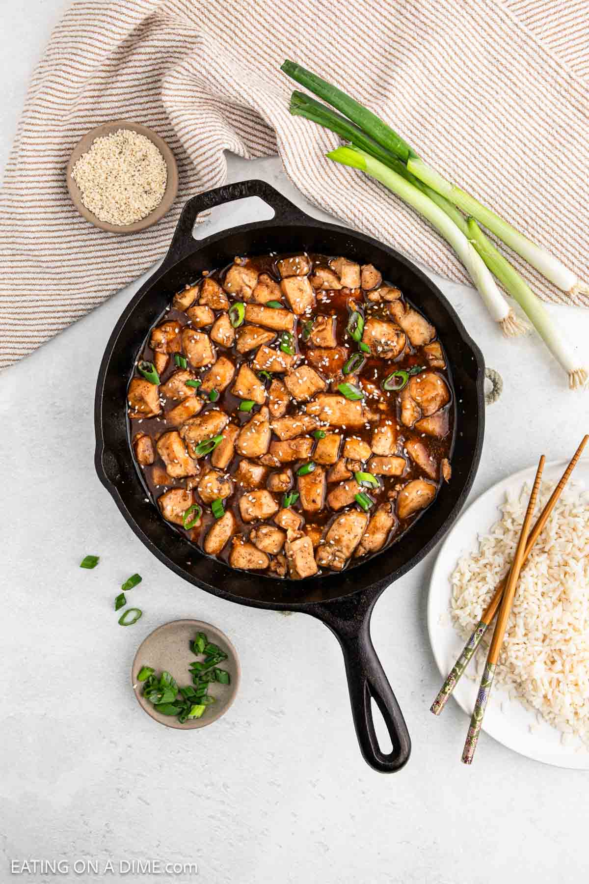 A skillet filled with Hoisin Chicken in a rich brown sauce, garnished with sesame seeds and green onions. Nearby, a bowl of rice with chopsticks, a small dish of sliced green onions, a bundle of fresh green onions, and sesame seeds on a cloth.