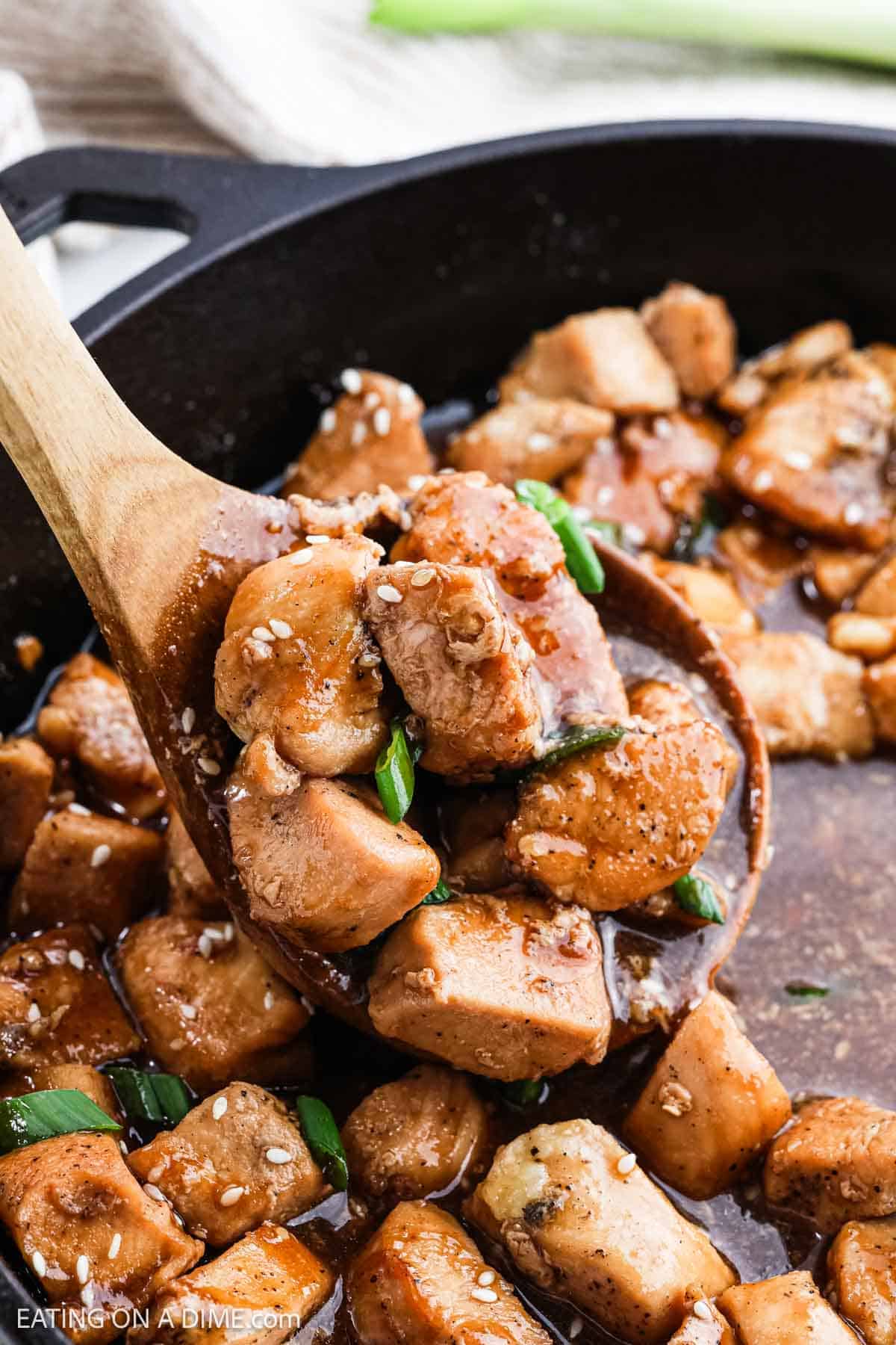 A skillet filled with Hoisin Chicken features cooked pieces coated in a rich, glossy sauce. A wooden spoon scoops up some chicken, garnished with sesame seeds and chopped green onions.