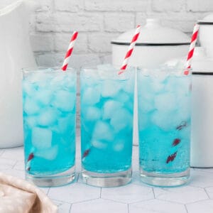 Three tall glasses filled with a vibrant blue liquid and ice cubes, reminiscent of an easy Sonic Ocean Water recipe, each adorned with a red and white striped straw. They rest on a hexagonal-tiled surface, with white kitchen containers gracefully positioned in the background.