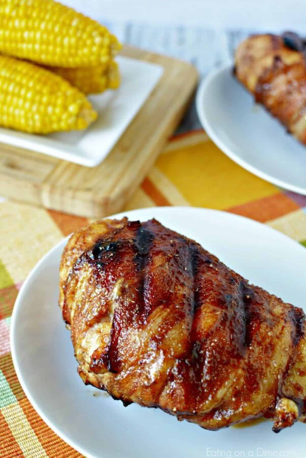 Bacon wrapped chicken breast on a plate with corn on the cob on a plate in the background