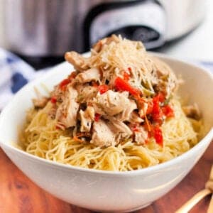 A bowl filled with cooked pasta topped with shredded chicken, red bell peppers, and grated cheese sits enticingly on a wooden surface. Reminiscent of an easy Chicken Scampi recipe, the flavors meld beautifully against the blurred kitchen appliance backdrop.