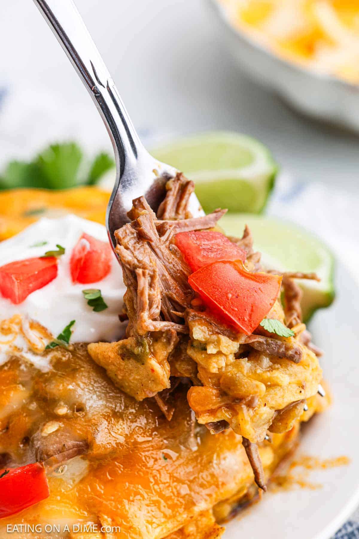 A serving of the beef enchiladas on a plate topped with sour cream and diced tomatoes with a bite on a fork