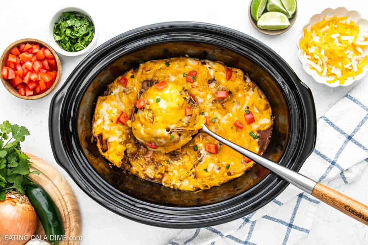 Beef enchiladas in the slow cooker topped with melted cheese and diced tomatoes with a spatula on top with small bowls of diced tomatoes, shredded cheese and fresh cilantro on the side