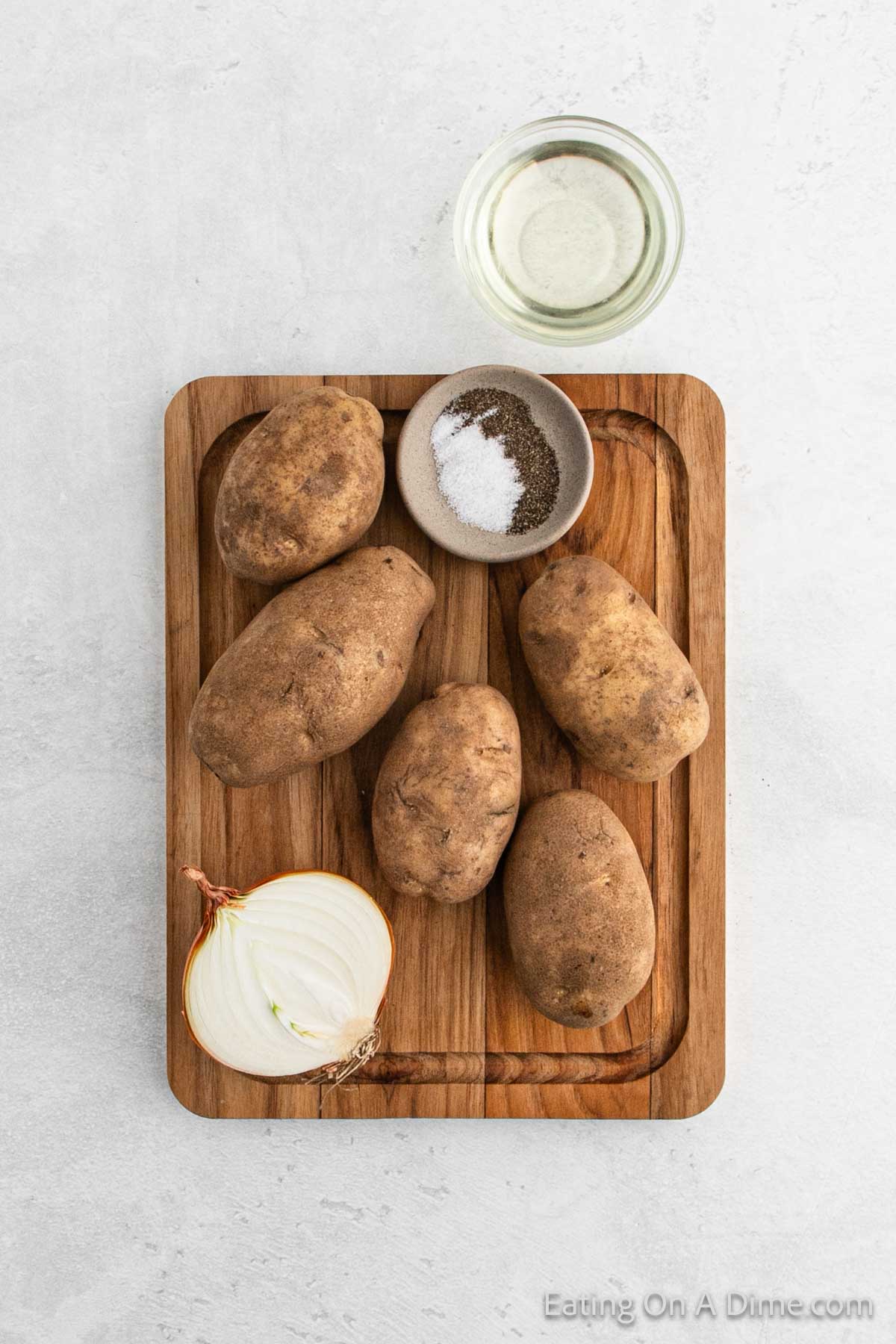 Five whole potatoes and half an onion rest on a wooden cutting board, ready to be transformed into delicious pan-fried potatoes. A small bowl of salt and pepper, along with a glass of oil, waits nearby to add flavor to the perfect crispy dish.