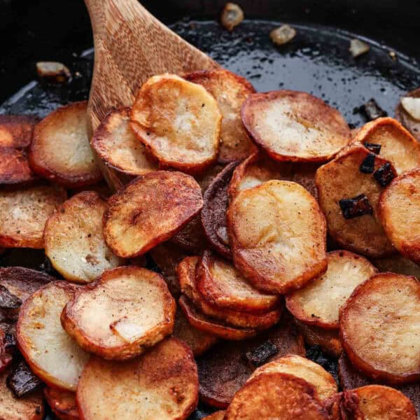 Close-up of golden-brown pan fried potatoes in a skillet, being stirred with a wooden spoon. The potatoes are crispy and sprinkled with small bits of cooked onion, showcasing a savory, home-cooked dish.