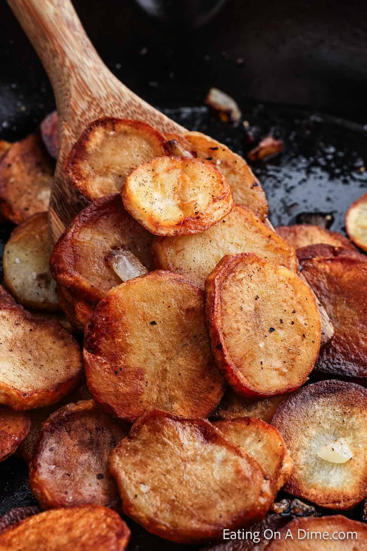 A close-up of a wooden spoon scooping Pan Fried Potatoes from a black skillet. The golden-brown potatoes are seasoned with visible pepper and small onion pieces, highlighting their crispy texture.