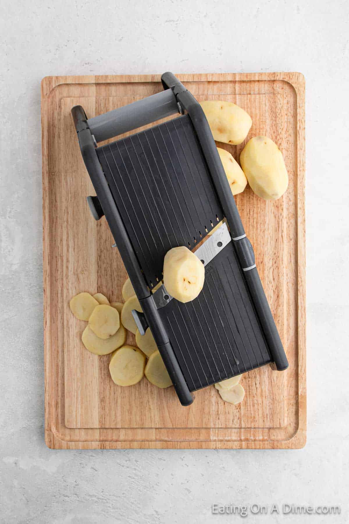 A mandoline slicer sits on a wooden cutting board with peeled potatoes, preparing for delicious pan-fried potatoes. Several slices are scattered around, and one potato is poised on the slicer, ready for its turn in the skillet.