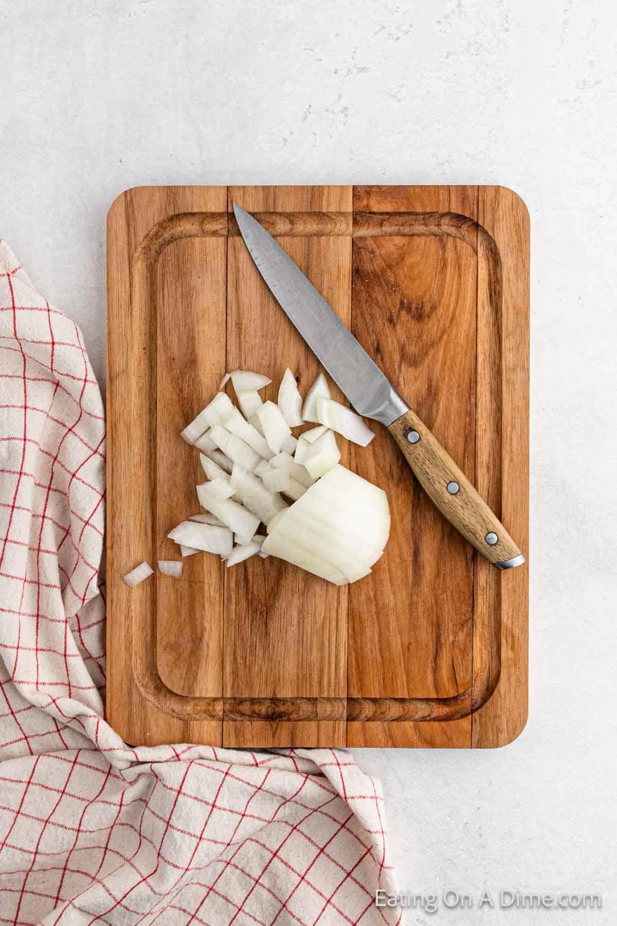 A wooden cutting board with chopped white onion and a knife sits beside a sizzling pan of fried potatoes. A red and white checkered cloth rests on the light-colored countertop, completing the cozy kitchen scene.