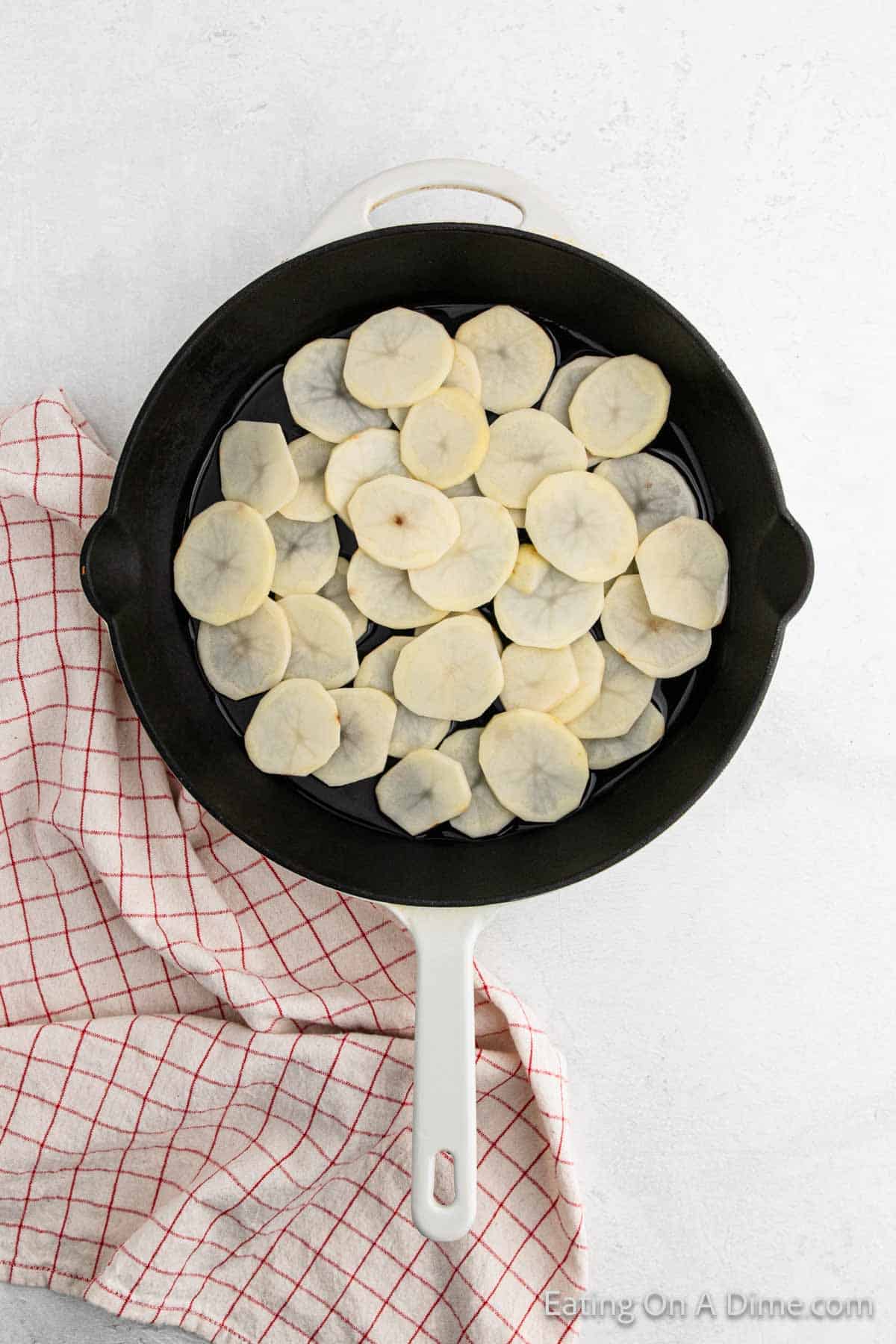 A white cast iron skillet filled with thin slices of taro root, reminiscent of pan-fried potatoes, rests on a light gray surface. A red and white checkered cloth is partially visible on the left side.