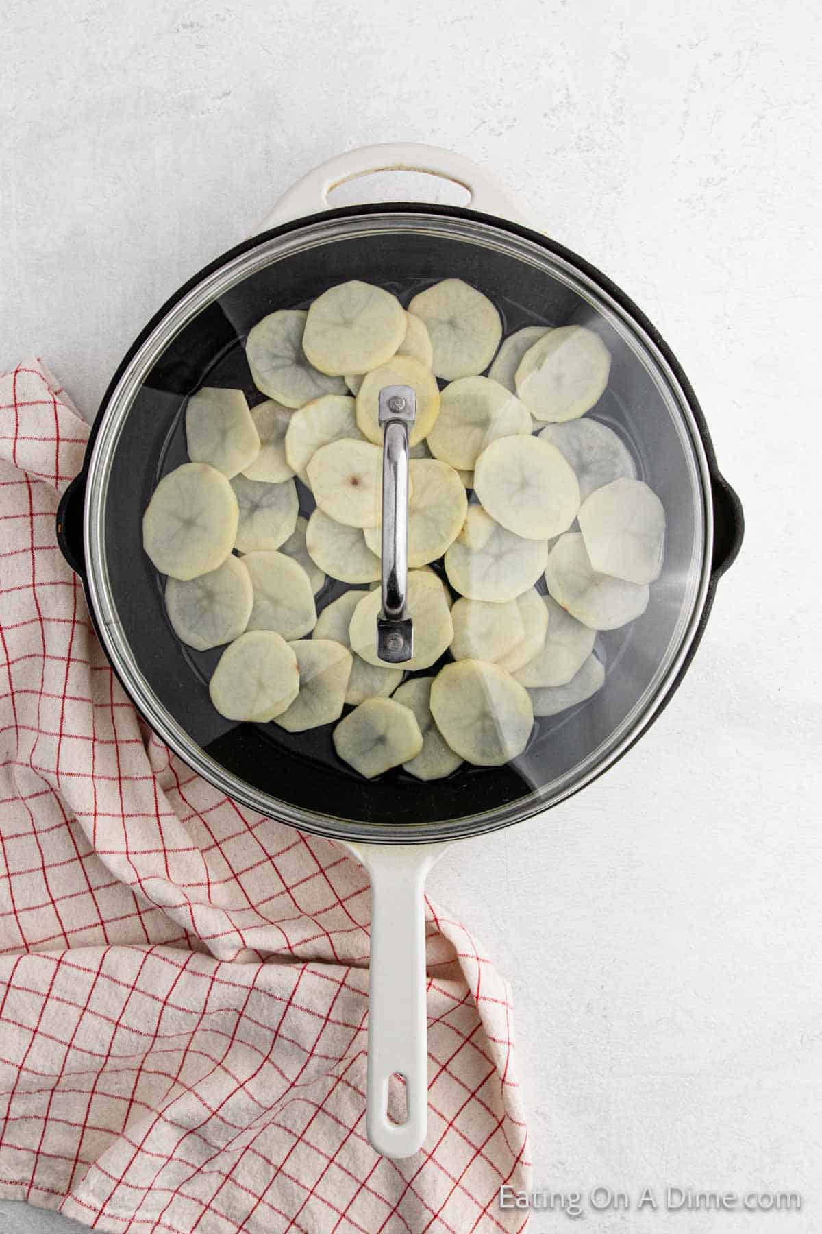 A white skillet with a glass lid, brimming with pan-fried potatoes, sits on a white surface. A red and white checkered towel lies beside the skillet, adding a touch of charm.