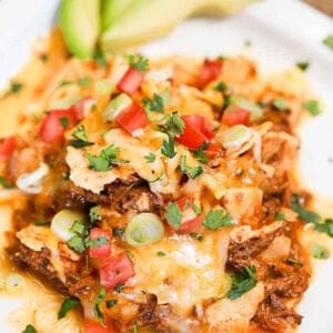 A plate of crock pot shredded beef enchilada casserole topped with melted cheese, chopped tomatoes, green onions, and fresh cilantro, with avocado slices in the background.