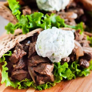 Close-up of a pita sandwich filled with grilled beef reminiscent of a crock pot beef gyros recipe, fresh green lettuce, and topped with a dollop of creamy white tzatziki sauce, served on a wooden surface.