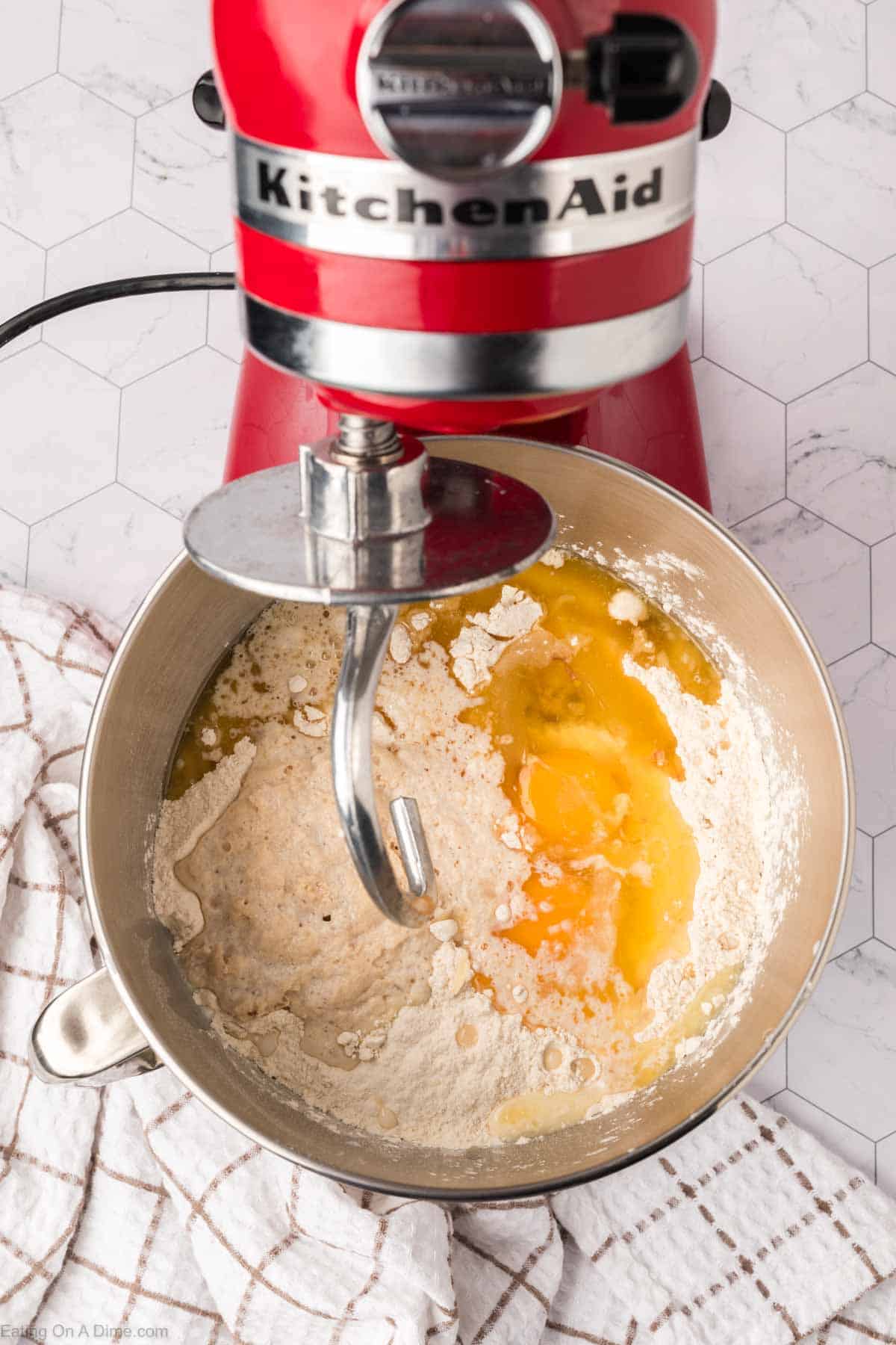 A red stand mixer with a dough hook attachment is vigorously blending ingredients for Mexican sweet bread. The bowl contains flour, eggs, and water. Nearby, a white and brown checkered towel rests on a hexagonal patterned countertop.