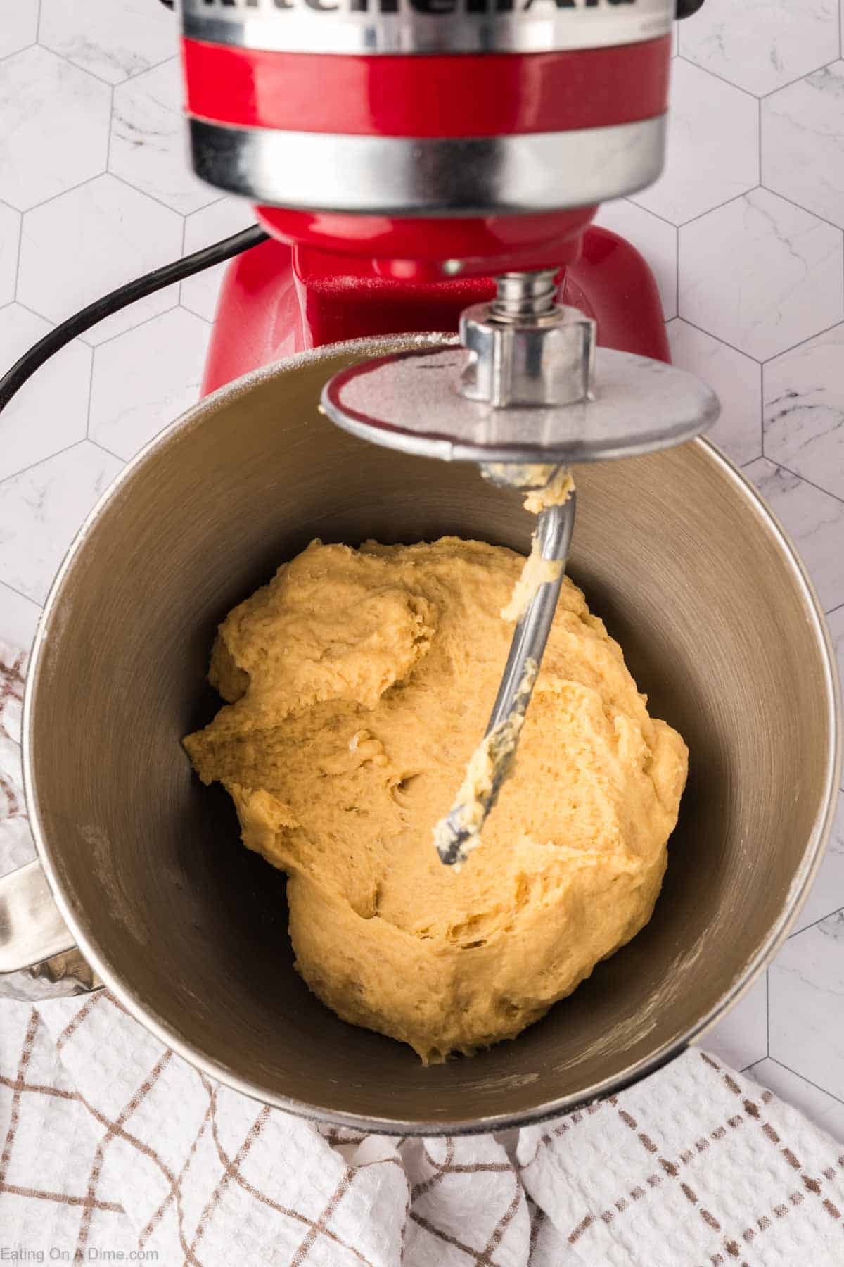 A red stand mixer with a metal bowl containing dough for Mexican sweet bread is in use. The dough, slowly transforming under the dough hook attachment, rests on a hexagon-patterned surface with a checkered cloth beneath.