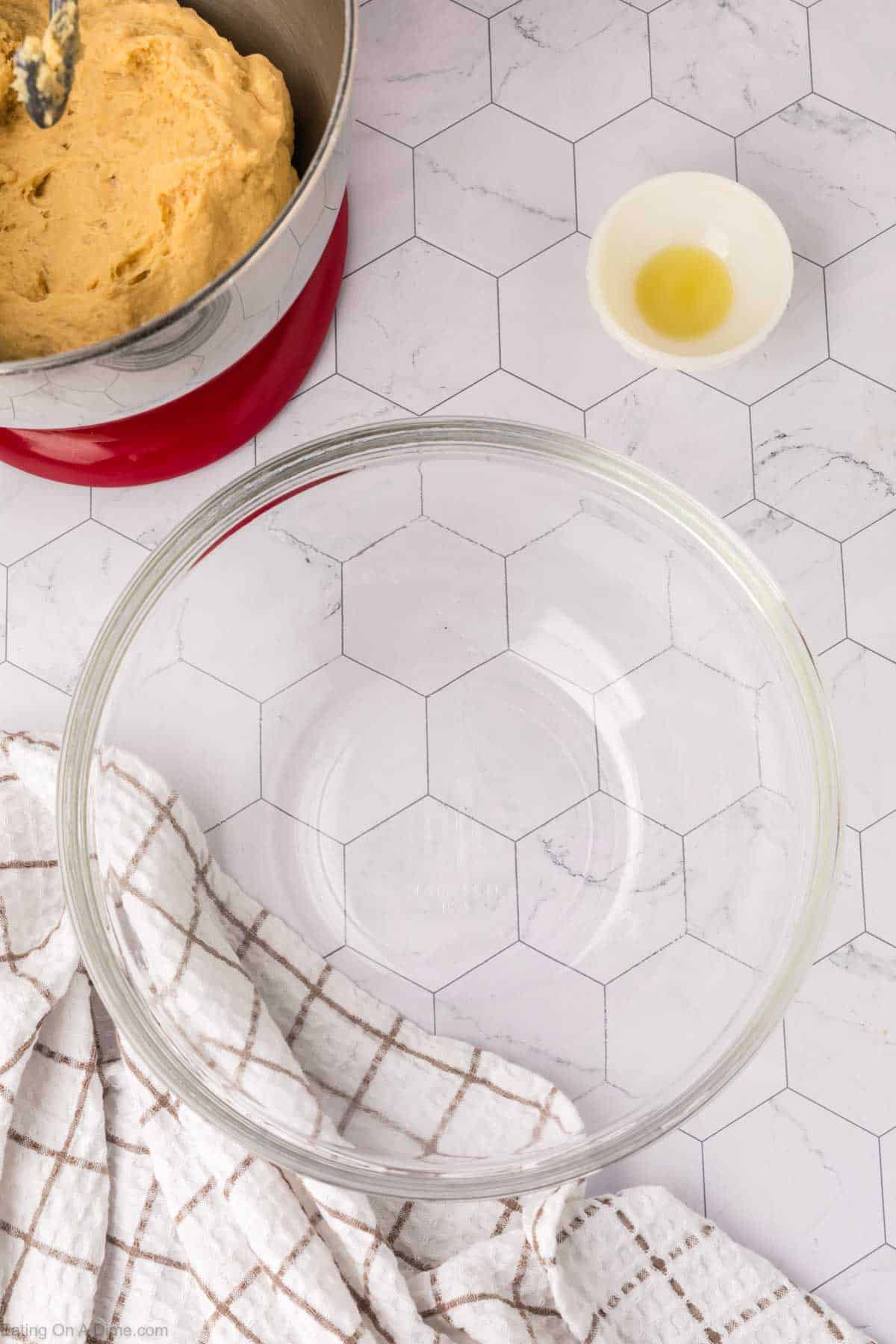 A red mixing bowl with cookie dough, reminiscent of Mexican sweet bread, sits alongside a small bowl with vanilla extract and a large empty glass bowl, all on a hexagonal-patterned countertop. A white kitchen towel with a brown grid pattern is partly draped over the counter.