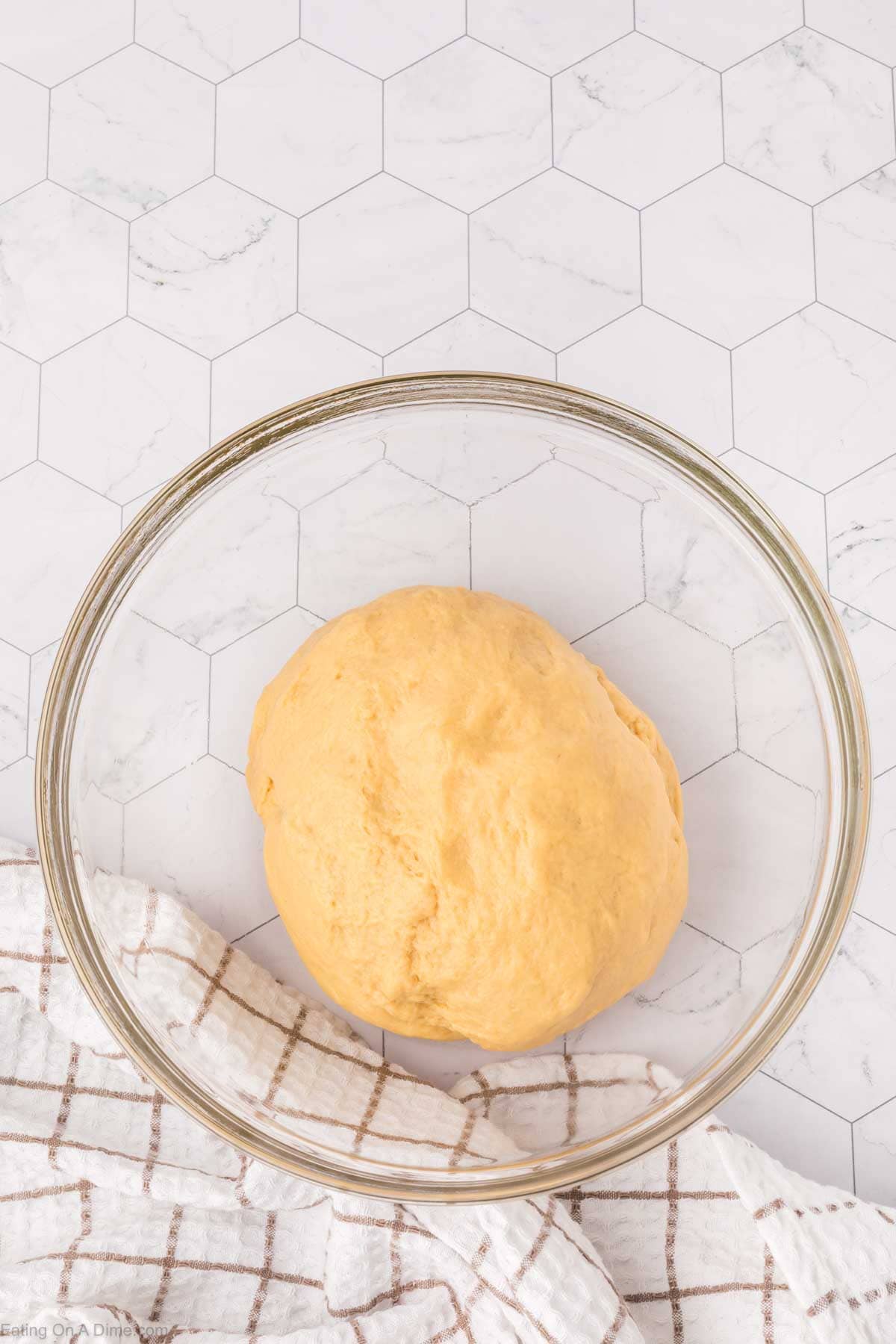 A ball of yellow dough, reminiscent of Mexican sweet bread, rests in a transparent glass bowl on a hexagonal-tiled countertop. A white cloth with a brown checkered pattern is partially visible at the bottom left corner.