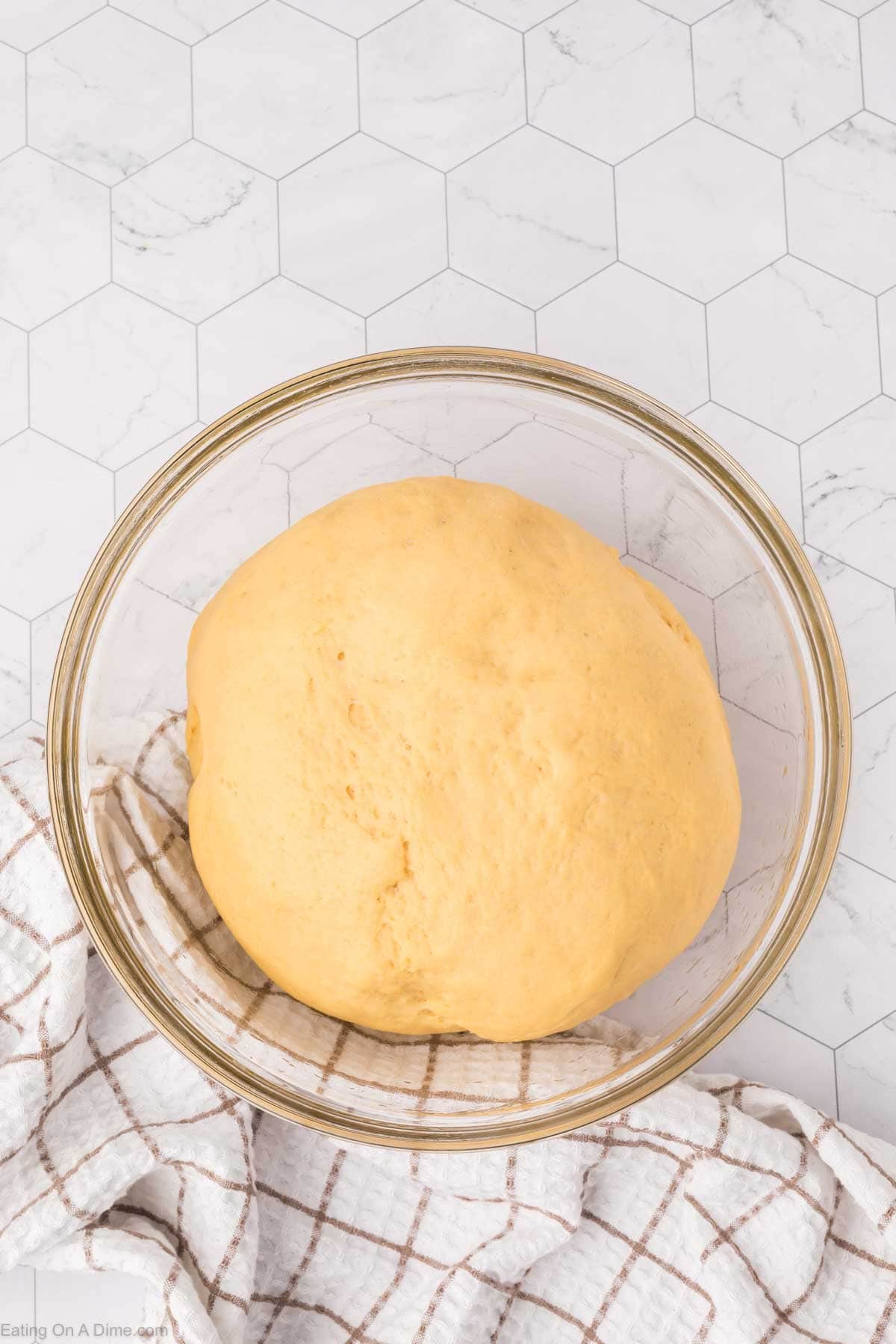 A ball of dough, destined for Mexican sweet bread, rests in a clear glass bowl on a white hexagonal-patterned surface. A checkered cloth is partially visible in the foreground.