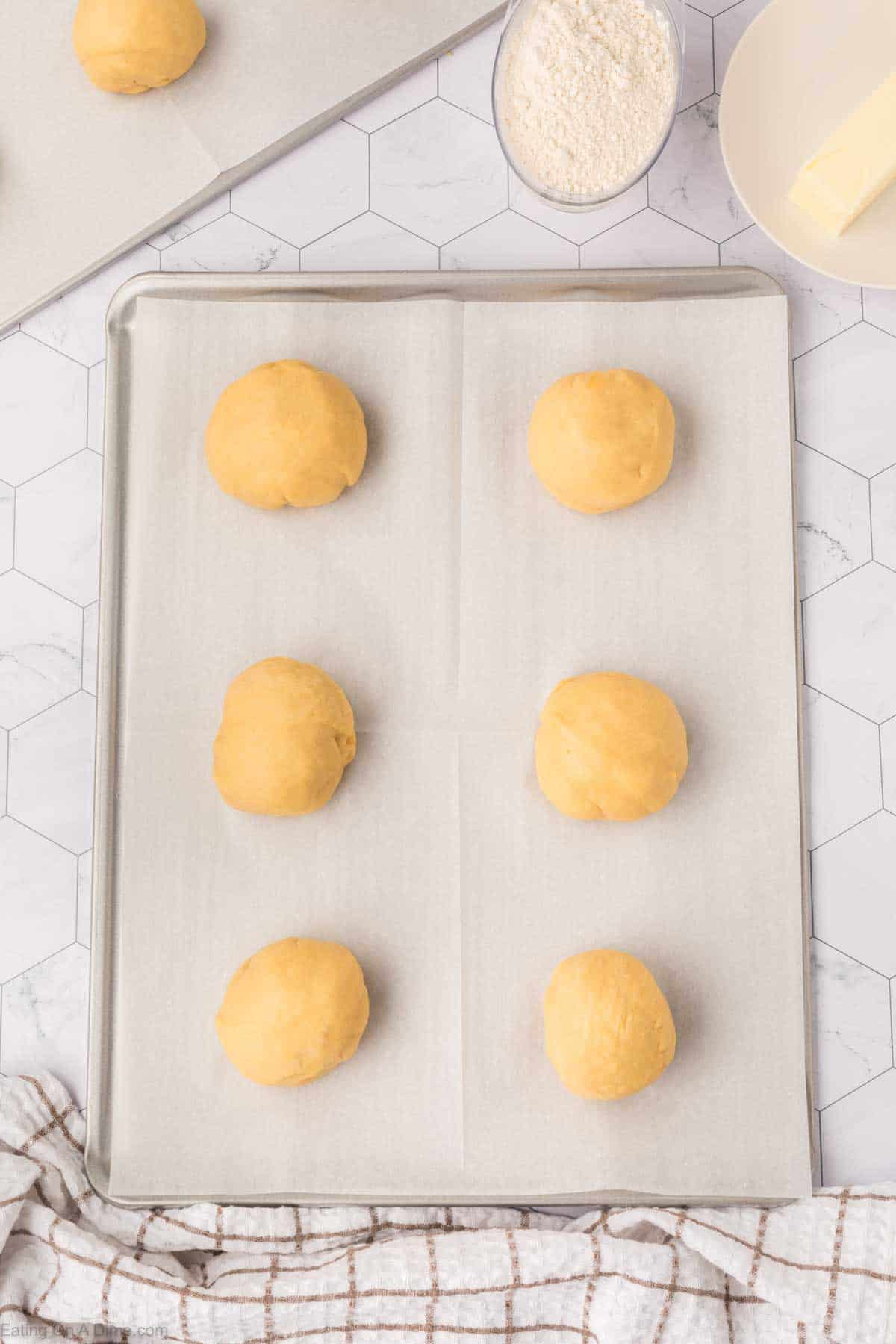 Six evenly spaced dough balls, soon to become Mexican sweet bread, rest on a parchment-lined baking sheet. Nearby are a bowl of flour and a small plate with butter. A checkered cloth graces the foreground against the backdrop of a hexagonal tile surface.