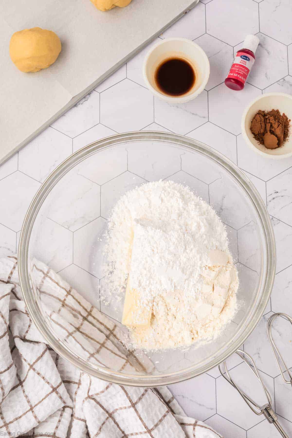 A glass bowl containing flour and butter sits on a kitchen counter, hinting at the making of Mexican Sweet Bread. Nearby are a small bowl of vanilla extract, cocoa powder, a red food coloring bottle, two dough balls, and a whisk. A checkered cloth lies beside them.