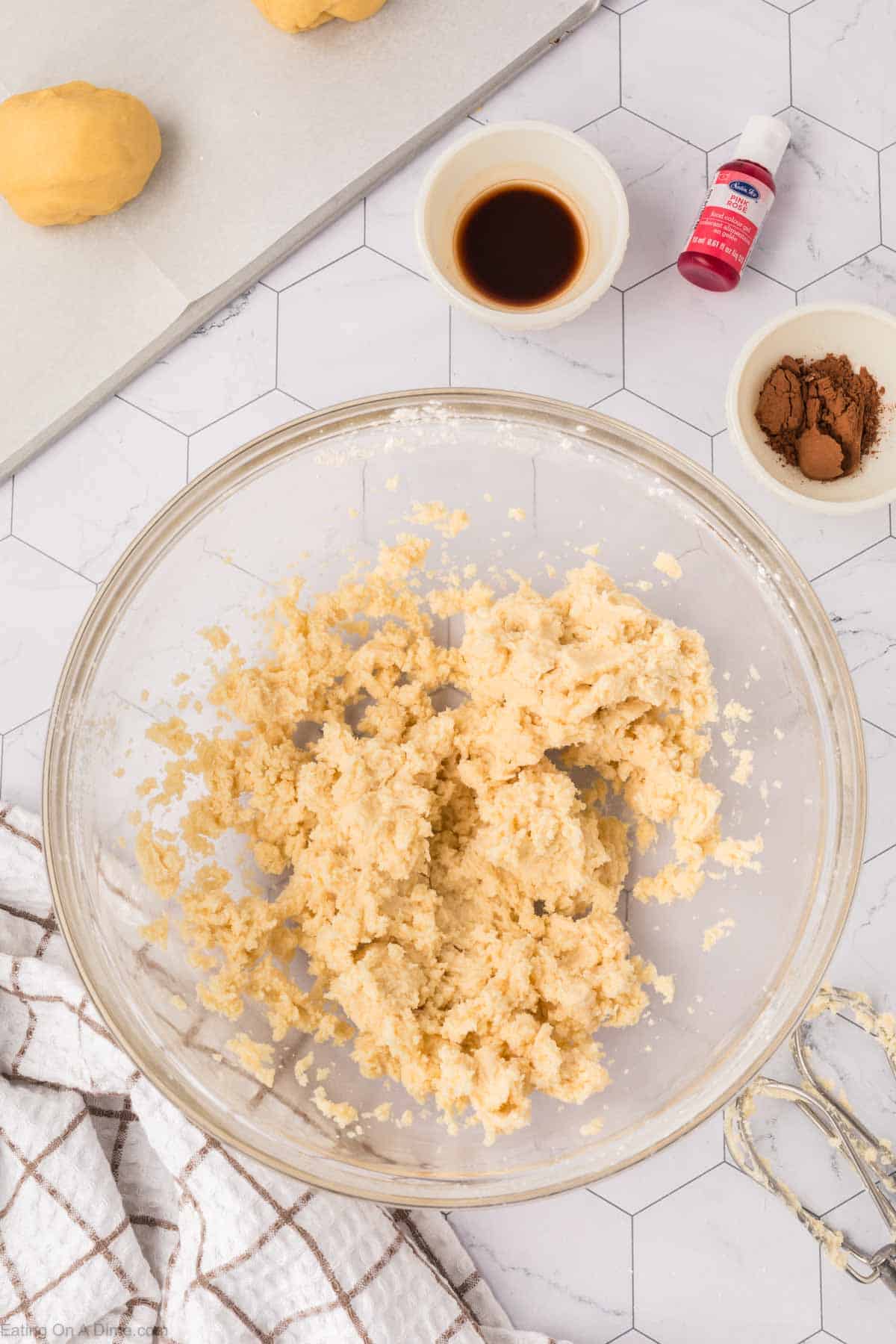 A glass bowl with cookie dough sits on a hexagonal patterned countertop, echoing the artful design found in Mexican Sweet Bread. Nearby are small bowls containing cocoa powder and a brown liquid, a red food coloring bottle, and cookie dough pieces on a cutting board. A plaid towel is partially visible.