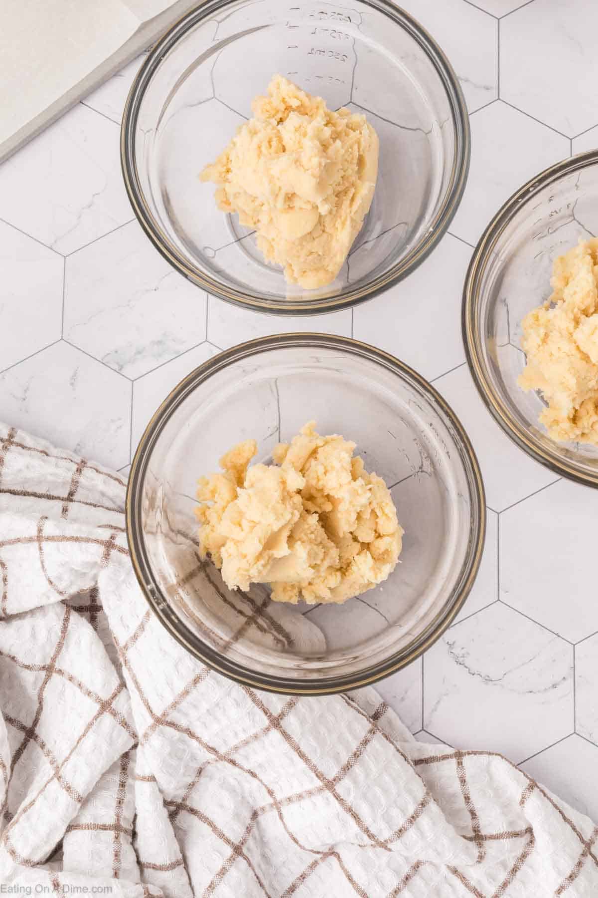 Three glass bowls with portions of dough, perhaps destined to become Mexican sweet bread, are placed on a white hexagonal tile surface. A white and brown checkered cloth is draped on the left side.