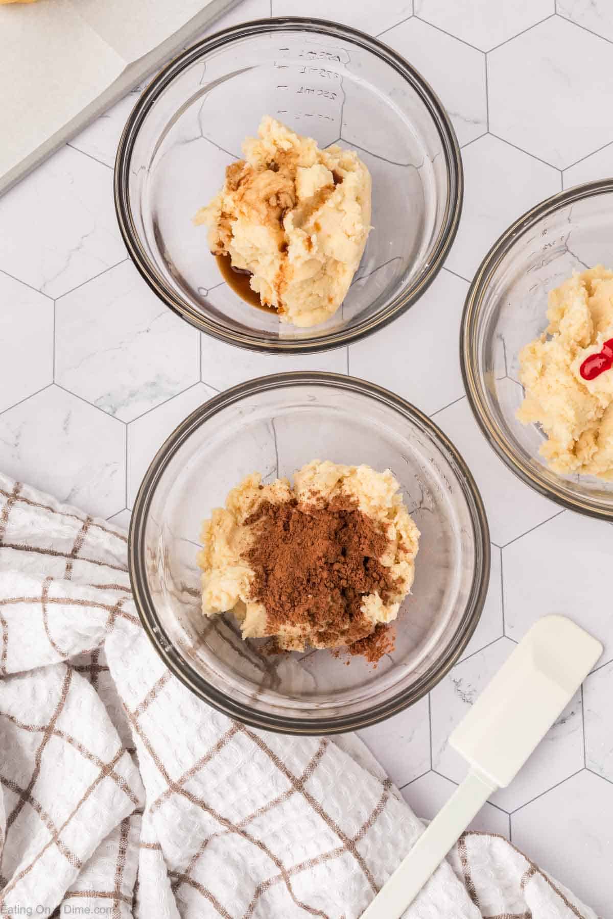 Three glass bowls on a white hexagon-patterned surface contain cookie dough mixtures reminiscent of Mexican sweet bread. One has vanilla extract, another cocoa powder, and the third red food coloring. A white spatula and a checkered towel are beside the bowls.