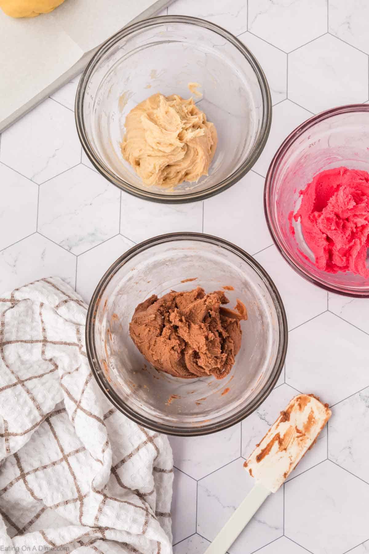 Three glass bowls display doughs in light brown, dark brown, and pink hues, reminiscent of the vibrant colors of Mexican Sweet Bread. Nearby, a white and brown striped cloth accompanies a cream-colored spatula with traces of brown dough on its edge. All rest on a white hexagonal patterned tabletop.