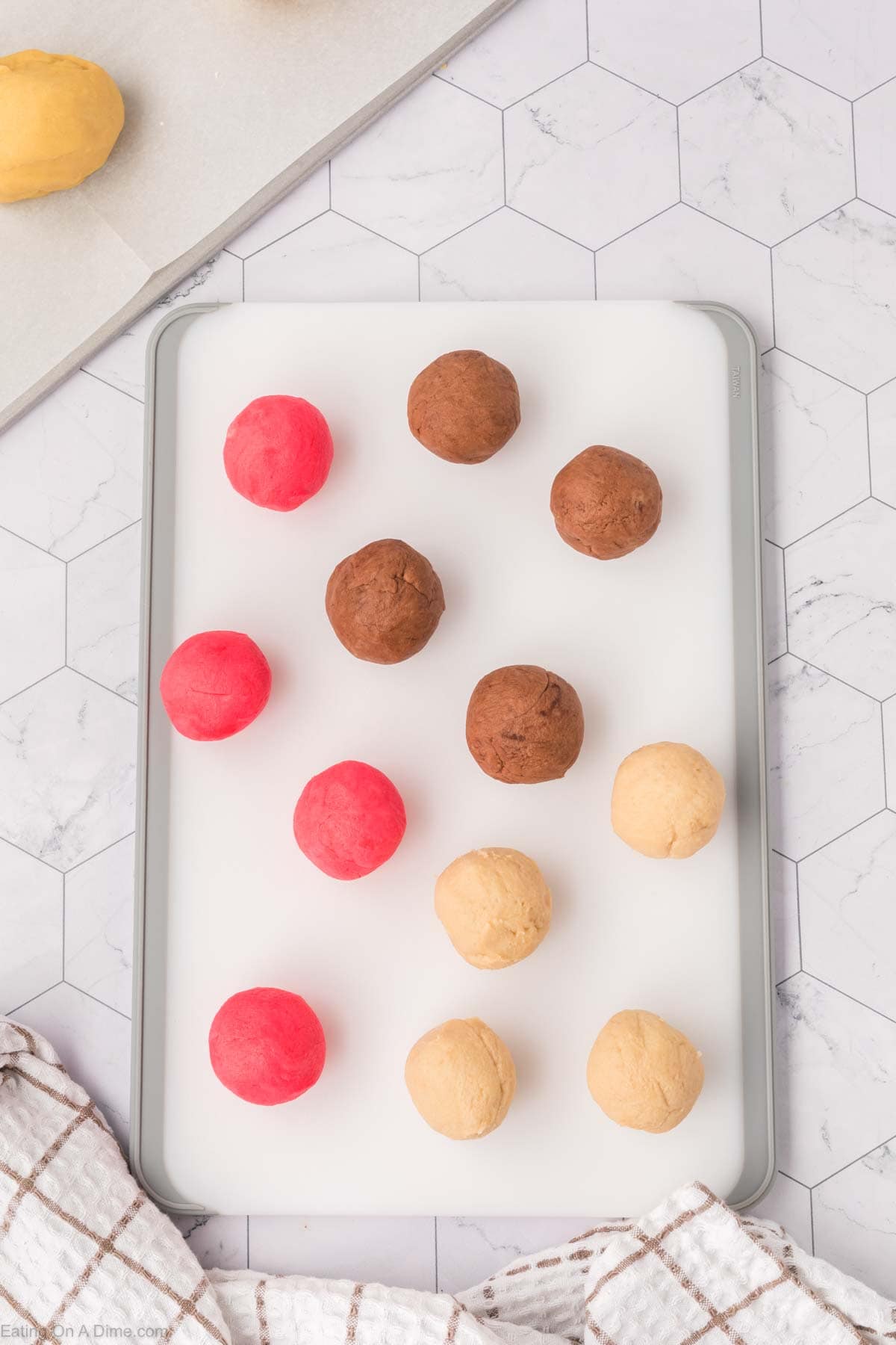 A baking tray with cookie dough balls, reminiscent of Mexican Sweet Bread, features three red, three brown, and three beige balls. The tray rests on a white hexagonal tiled surface, with a kitchen towel partially visible in the bottom corner.