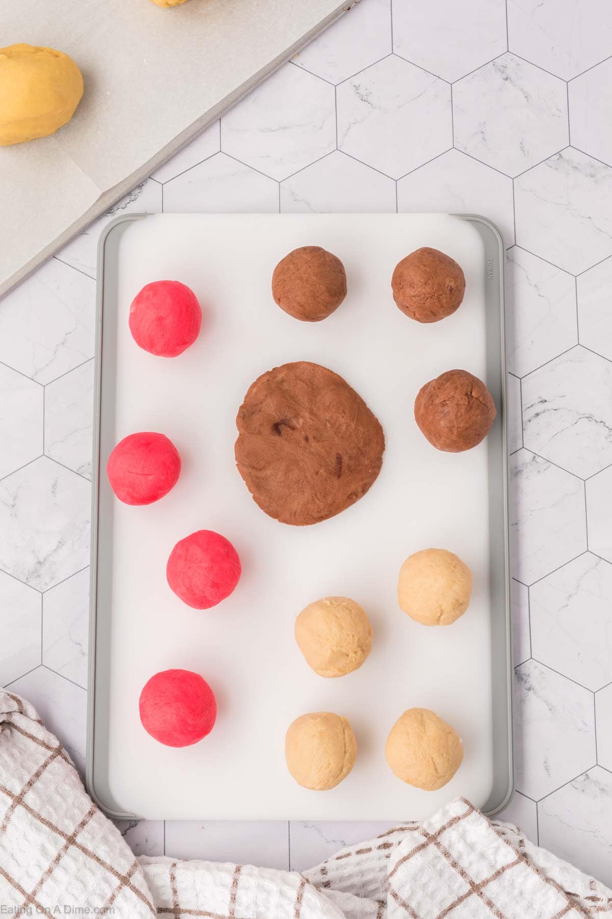 Balls of red, brown, and beige dough reminiscent of Mexican Sweet Bread are arranged on a white cutting board. A large flattened piece of brown dough rests in the center. A plaid cloth peeks into view at the bottom left corner.