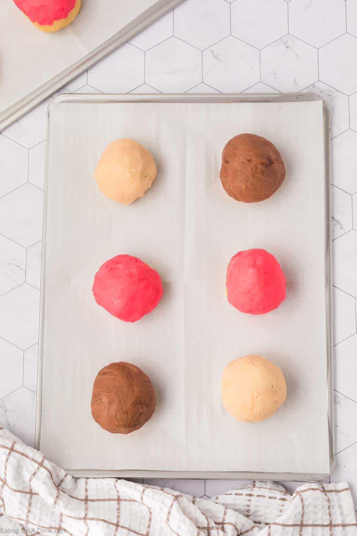 Six colored dough balls, reminiscent of Mexican sweet bread, sit evenly spaced on a parchment-lined baking sheet. Included are two red, two brown, and two beige balls. A hexagon-patterned surface is visible underneath, alongside part of a striped cloth.