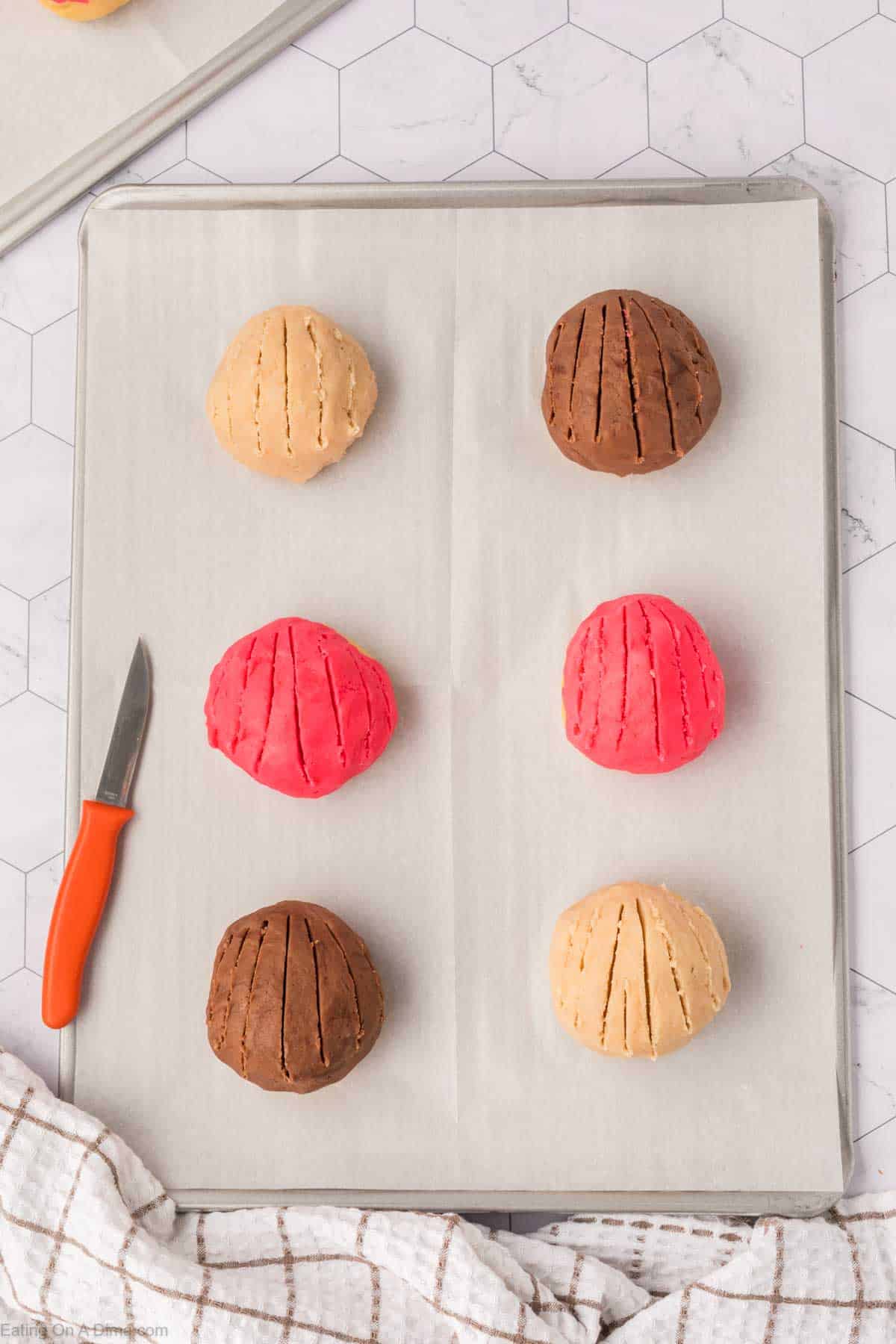 Six dough balls, reminiscent of Mexican Sweet Bread, three light brown and three pink, rest on a parchment-lined baking sheet. Each has three shallow cuts on top. A small knife with an orange handle lies to the left on a surface adorned with a white hexagonal tile design.