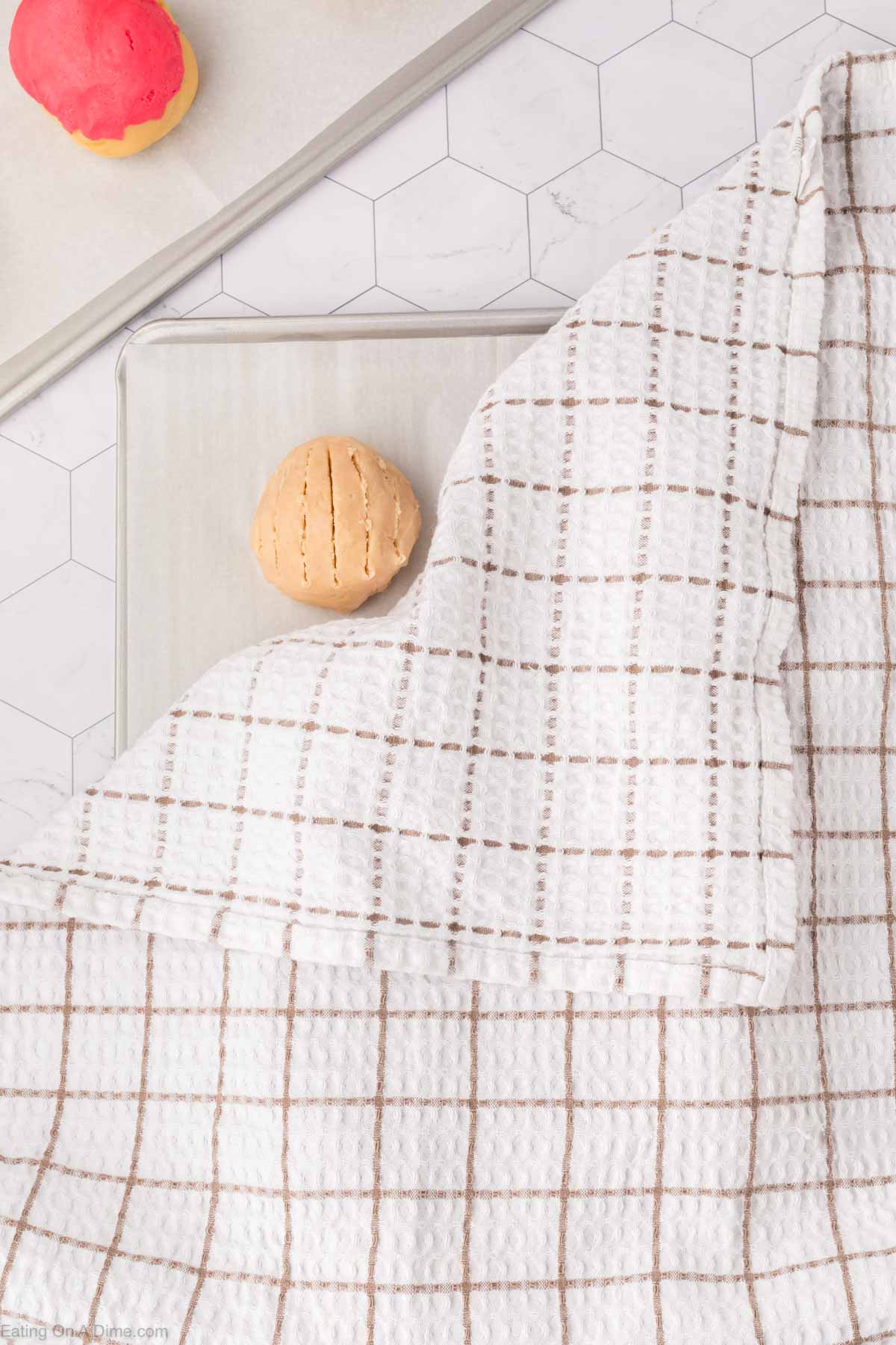 A ball of dough, reminiscent of Mexican sweet bread, rests on a countertop partially covered by a white checkered cloth. In the background, a baking sheet cradles a vibrant red and yellow dough ball. The hexagonal pattern of the countertop adds a touch of elegance to the scene.