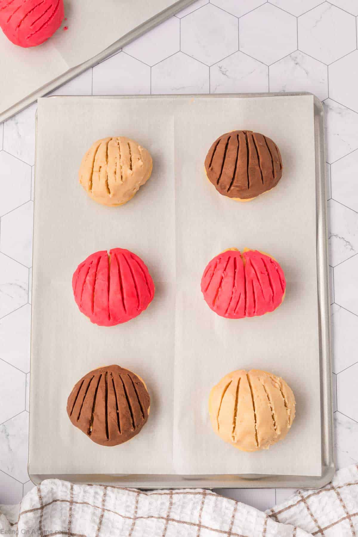 Six round, colorful pastries resembling Mexican Sweet Bread rest on a baking tray lined with parchment paper. With two red, two brown, and two beige delights—each boasting a scored top—they exude charm. A checkered cloth adds a cozy touch at the bottom edge of the image.