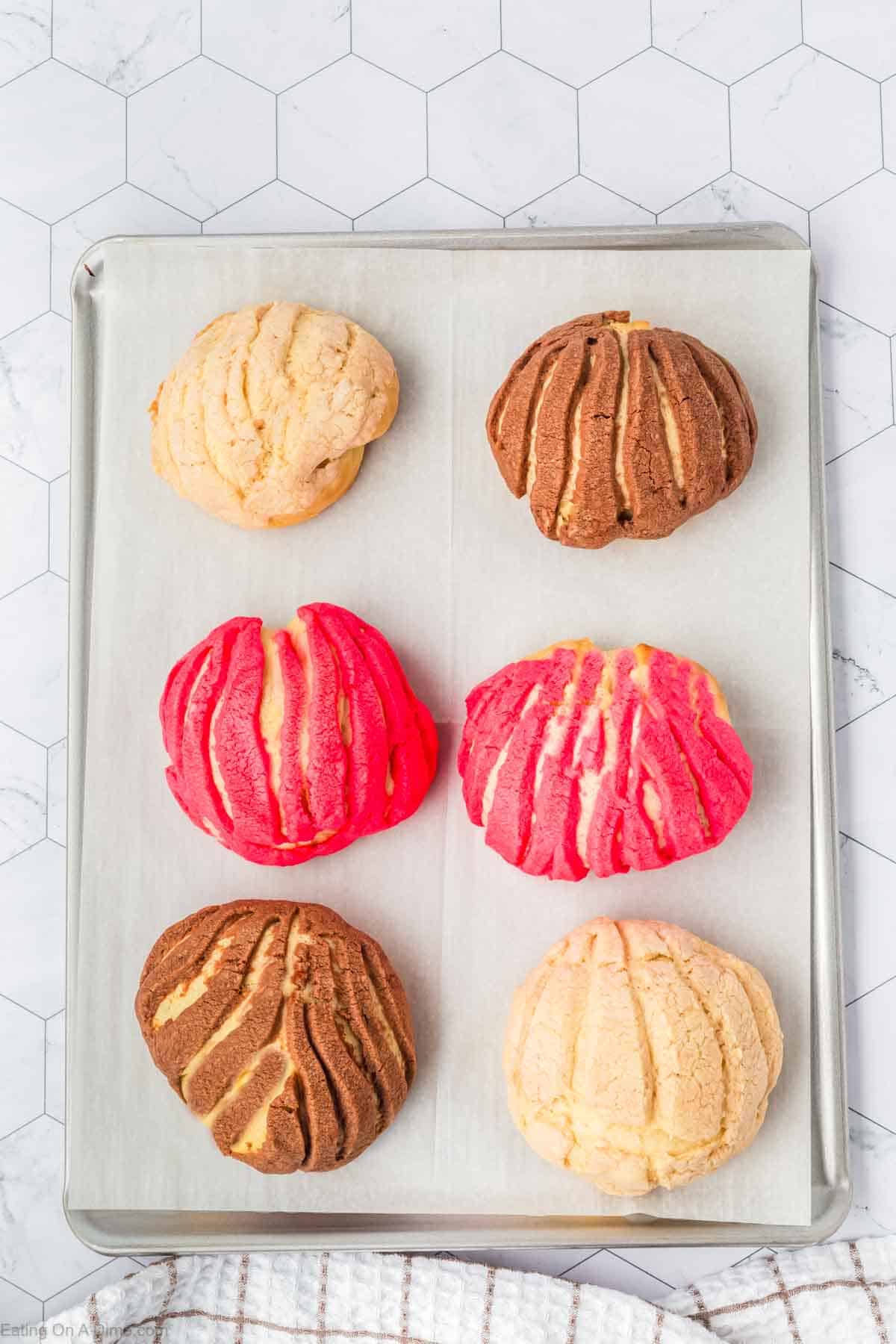 A baking tray with six Mexican sweet breads, or conchas, on parchment paper. The conchas feature colorful shells: two pink, two brown, and two beige. Their pattern resembles seashells. The tray rests on a white hexagonal patterned surface.