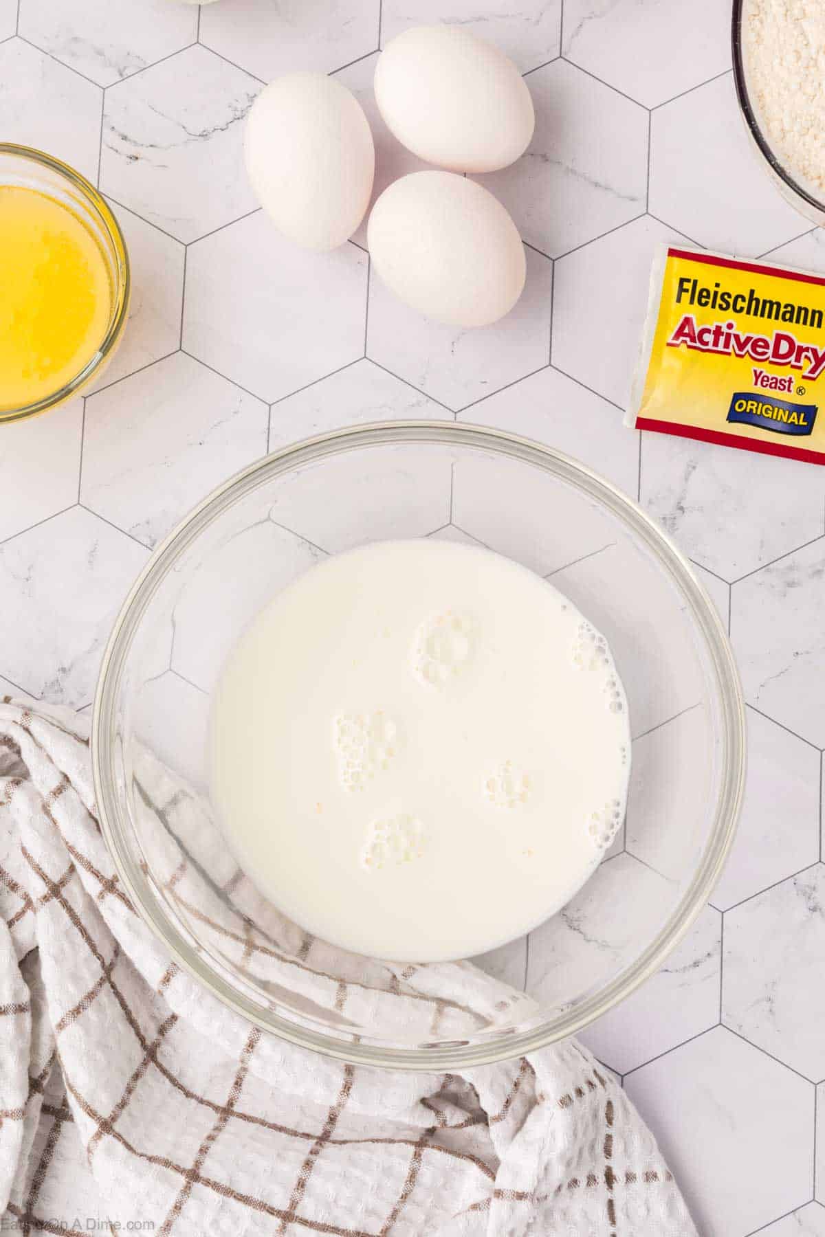 A glass bowl filled with milk sits on a hexagonal-patterned countertop, ready to be transformed into Mexican sweet bread. Nearby are three eggs, a packet of active dry yeast, a bowl of melted butter, and a patterned kitchen towel.