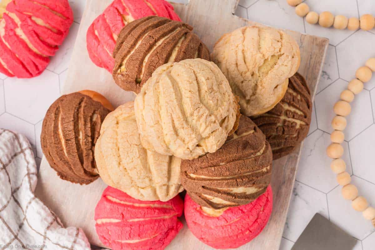 A pile of Mexican sweet bread, known as conchas, sits on a wooden board. The conchas are in three colors: pink, chocolate, and cream, each with a distinctive shell-like patterned topping. A hexagonal tile surface is underneath.