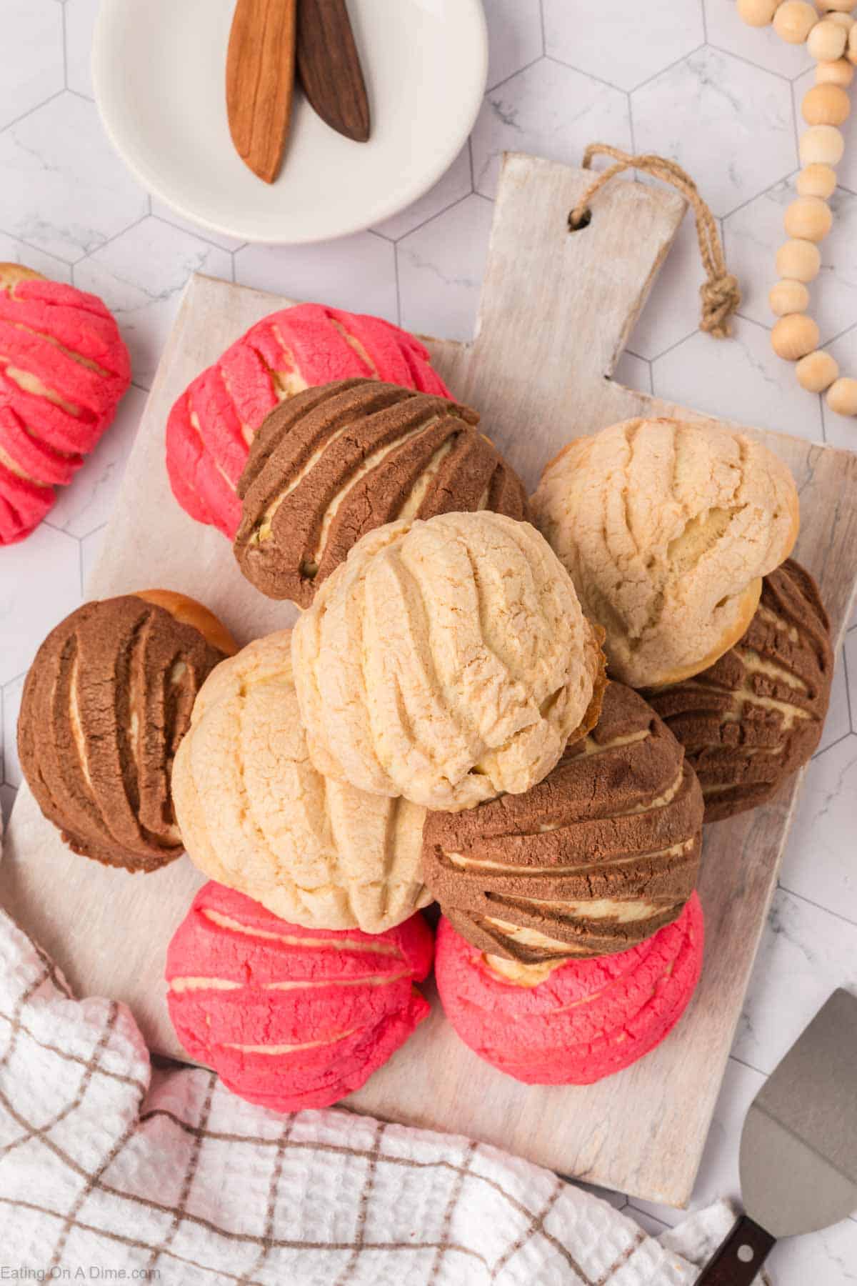 A stack of Mexican sweet breads, or conchas, in pink, chocolate, and vanilla flavors are artfully arranged on a wooden serving board. The background features a hexagonal tile pattern with a small plate and wooden spatulas nearby.