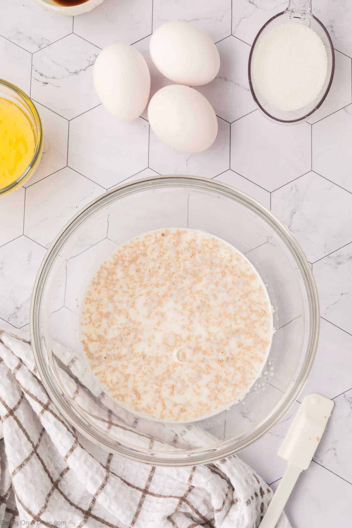 A glass bowl contains a mixture of liquid and yeast granules, hinting at the start of a Mexican sweet bread recipe. Surrounding the bowl are three eggs, a cup of sugar, a small bowl of melted butter, and a white checkered cloth on a hexagonal patterned surface.