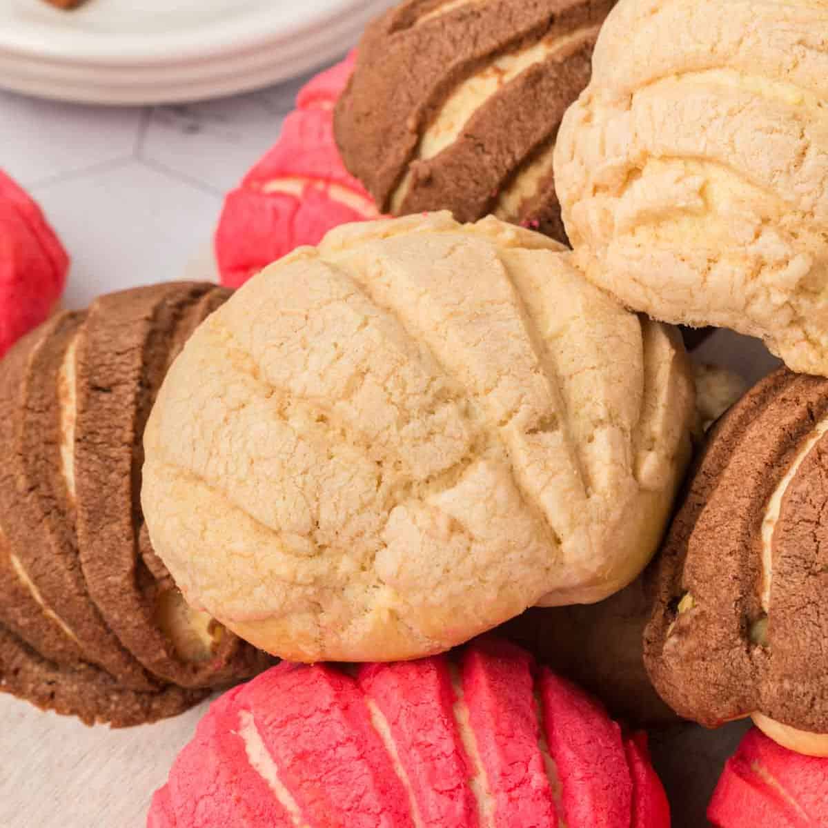 Close-up of assorted conchas, a type of Mexican sweet bread, on a light surface. They feature a shell-like pattern on top in pink, chocolate, and vanilla flavors. In the background, part of a white plate is visible.