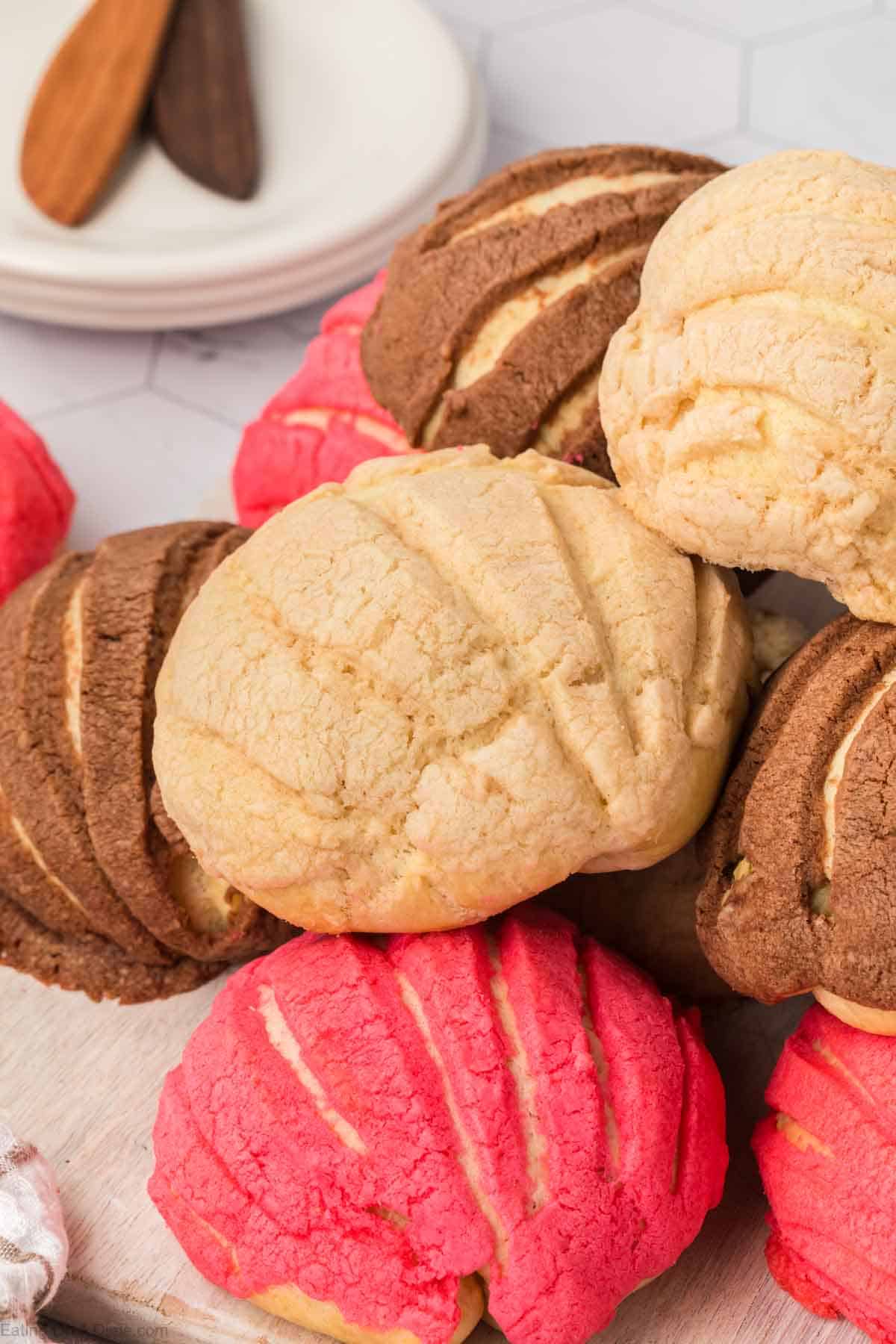 A close-up of a variety of conchas, Mexican sweet bread, showcases pink, brown, and beige sugar shell tops in a decorative pattern. In the background are two wooden butter knives and a stack of plates.