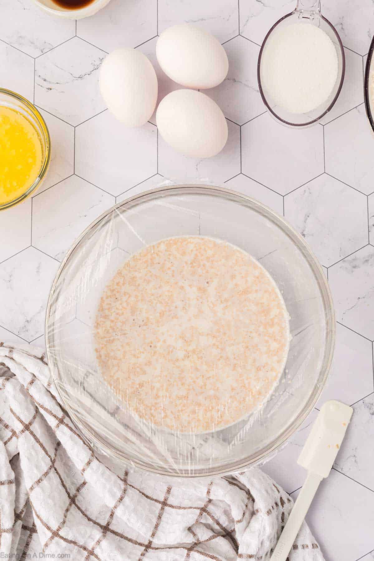 A bowl covered with plastic wrap, containing a liquid mixture for Mexican sweet bread, sits on a hexagonal patterned countertop. Surrounding it are glass bowls with melted butter and sugar, three eggs, a wooden spoon, and a white checkered towel.