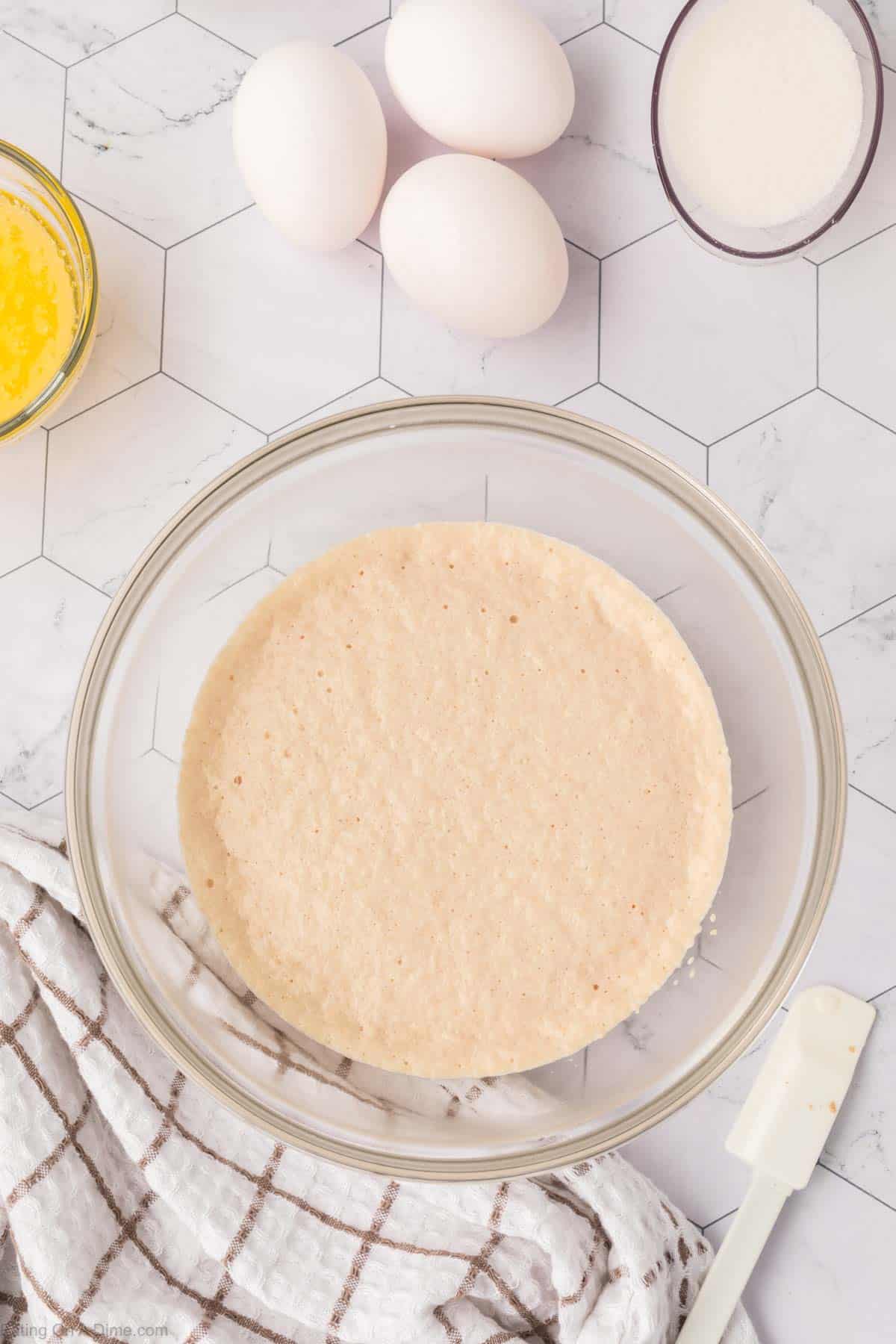 A glass bowl containing a light, thick batter for Mexican sweet bread sits on a hexagonal tiled surface. Nearby are three white eggs, a cup of sugar, melted butter, and a striped kitchen towel with a spatula.