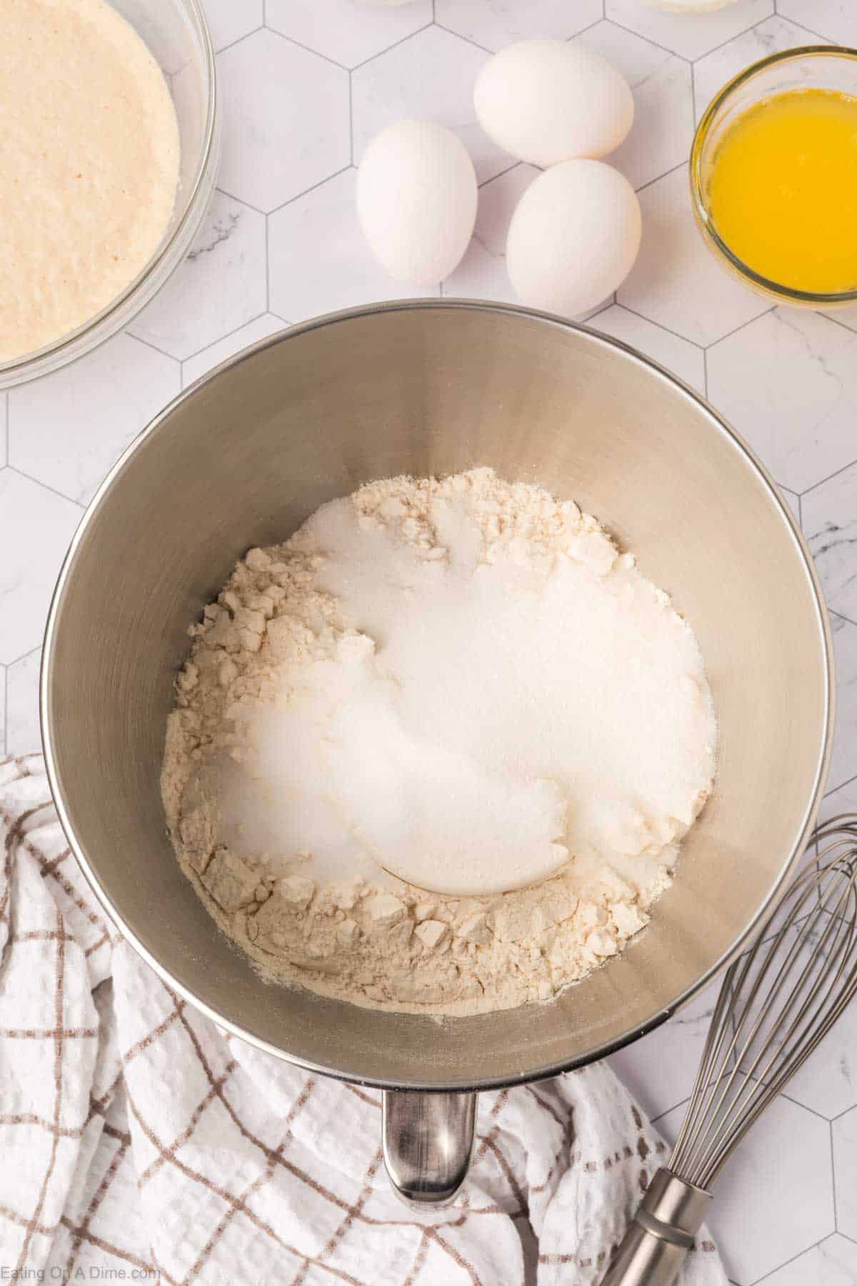 A mixing bowl containing flour and sugar sits on a hexagon-patterned countertop. Nearby are three eggs, a bowl of melted butter, and a towel, with a whisk ready to craft delicious Mexican Sweet Bread amidst bowls brimming with more ingredients.