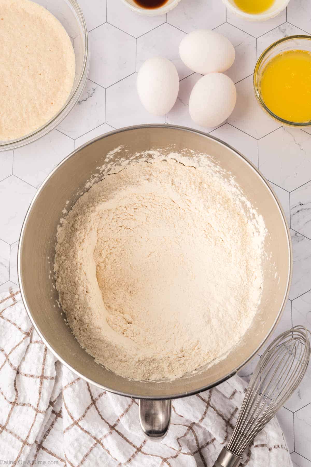 A mixing bowl filled with flour sits on a hexagonal-tiled surface, ready for Mexican sweet bread. Nearby are a whisk, a white and brown checkered towel, three eggs, a small bowl of melted butter, vanilla extract, and additional flour in a bowl.