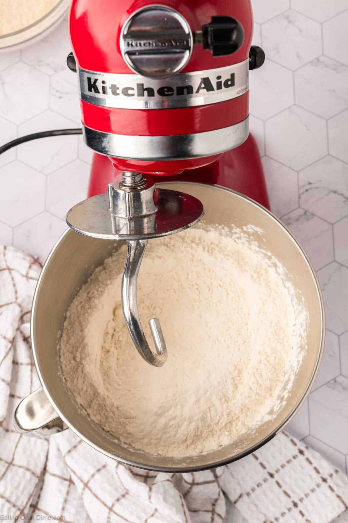 A red KitchenAid stand mixer with a dough hook attachment sits over a bowl filled with flour, ready to craft Mexican sweet bread. It's placed on a hexagonal tiled surface, accompanied by a white dish towel with a striped pattern nearby.